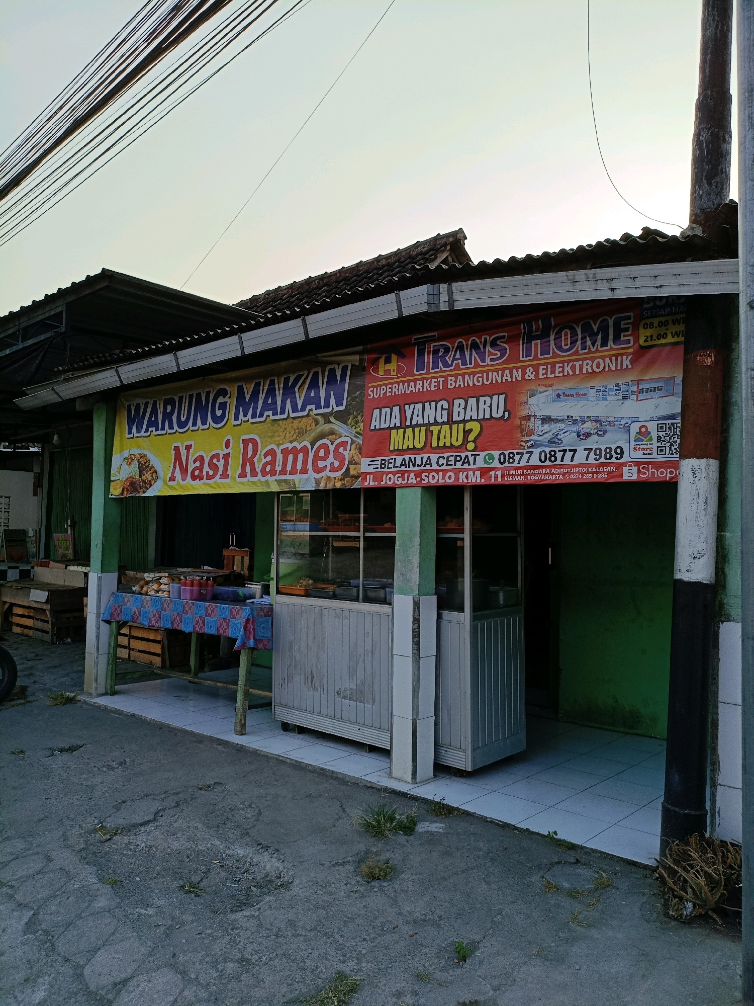 The image shows a storefront in Purwomartani, Indonesia, featuring two banners. The left banner, in yellow, reads "WARUNG MAKAN Nasi Rames" (Food Stall Mixed Rice) and displays an image of a plate of rice with various side dishes. Below this banner is a table covered with a patterned cloth, set up with food containers and condiments. The right banner is red and advertises "TRANS HOME SUPERMARKET BANGUNAN & ELEKTRONIK" (Trans Home Building & Electronics Supermarket) with the slogan "ADA YANG BARU, MAU TAU?" (Something New, Want to Know?). This banner also includes a phone number, address, and a QR code. The storefront has a green wall and a light-colored tiled floor. In front of the store is a grey concrete pavement with some damage and sparse vegetation. Electrical wires run overhead, and a utility pole is visible on the right side of the image. The overall scene depicts a commercial area with a mix of food and retail businesses.