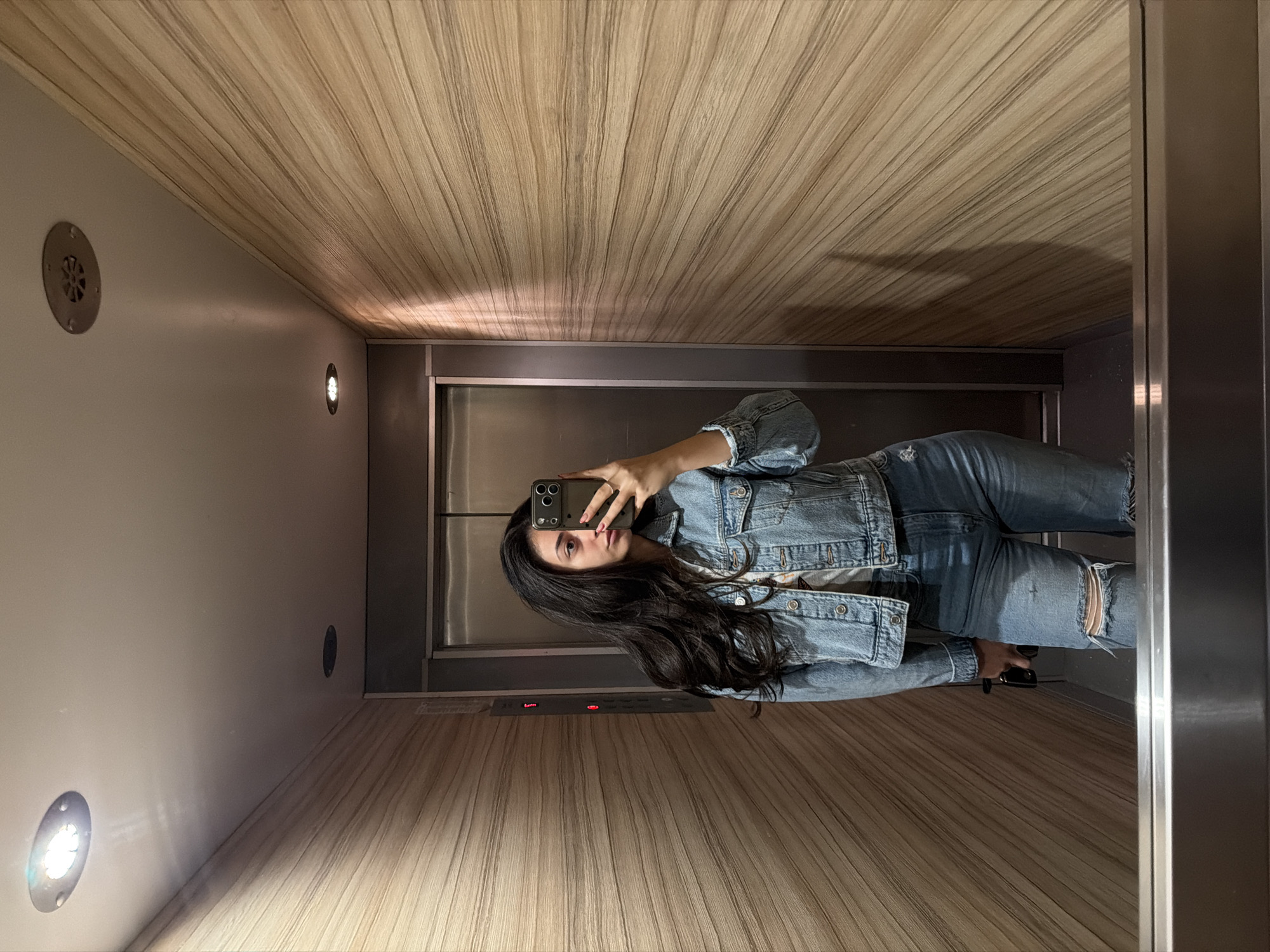 A woman is taking a selfie in an elevator. The interior of the elevator features light-colored wood paneling on the ceiling and one wall. Small, round lights are set into the wall opposite the wood paneling. There is a circular vent also on that wall. The floor is also covered in a light-colored wood-like material. The elevator has a metallic door with a control panel visible. The woman is wearing a denim jacket and jeans. The photograph does not provide any information that can be used to determine the location in Sofia, Bulgaria.