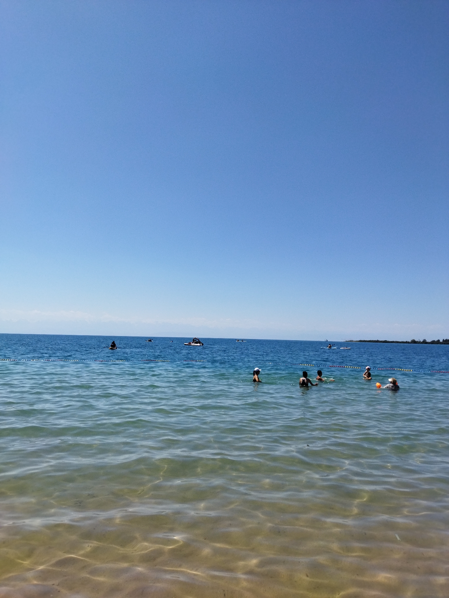 The image captures a sunny day at Lake Issyk-Kul in Kara-Oy, Kyrgyzstan. The clear blue sky stretches above a calm, blue body of water. In the foreground, the shallow water allows the sandy bottom to be visible, with gentle ripples reflecting the sunlight. Several people are enjoying the water. In the middle ground, closer to the shore, five individuals are partially submerged, some interacting with each other. Further out, several small boats and individuals on paddleboards are scattered across the lake. A yellow and blue rope barrier stretches across the water, marking a designated swimming area. The horizon shows a faint, hazy line of distant land, likely the other side of the lake.