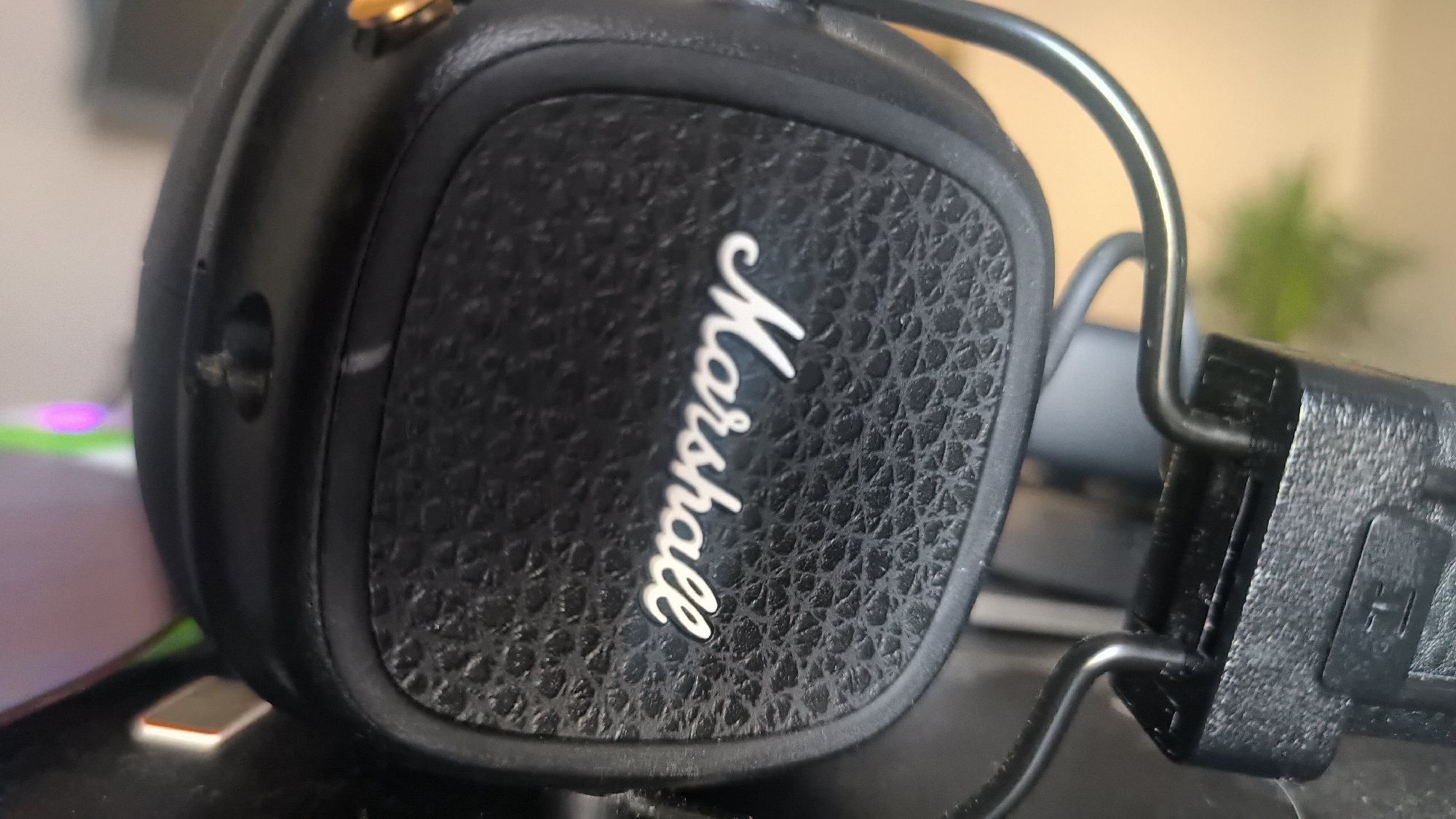 A close-up, side-angle shot focuses on a black Marshall brand headphone. The headphone features a textured, faux-leather earpad with the white "Marshall" logo embossed on it. A metal band, part of the headphone's headband, curves over the earpad. To the left of the earpad, a small black button or port is visible on the side of the headphone housing. The background is blurred, showing hints of a light-colored surface and a green plant. The image appears to be taken indoors, on a desk or table. There is no information to suggest this image is related to the city of Kovrov, Russia.