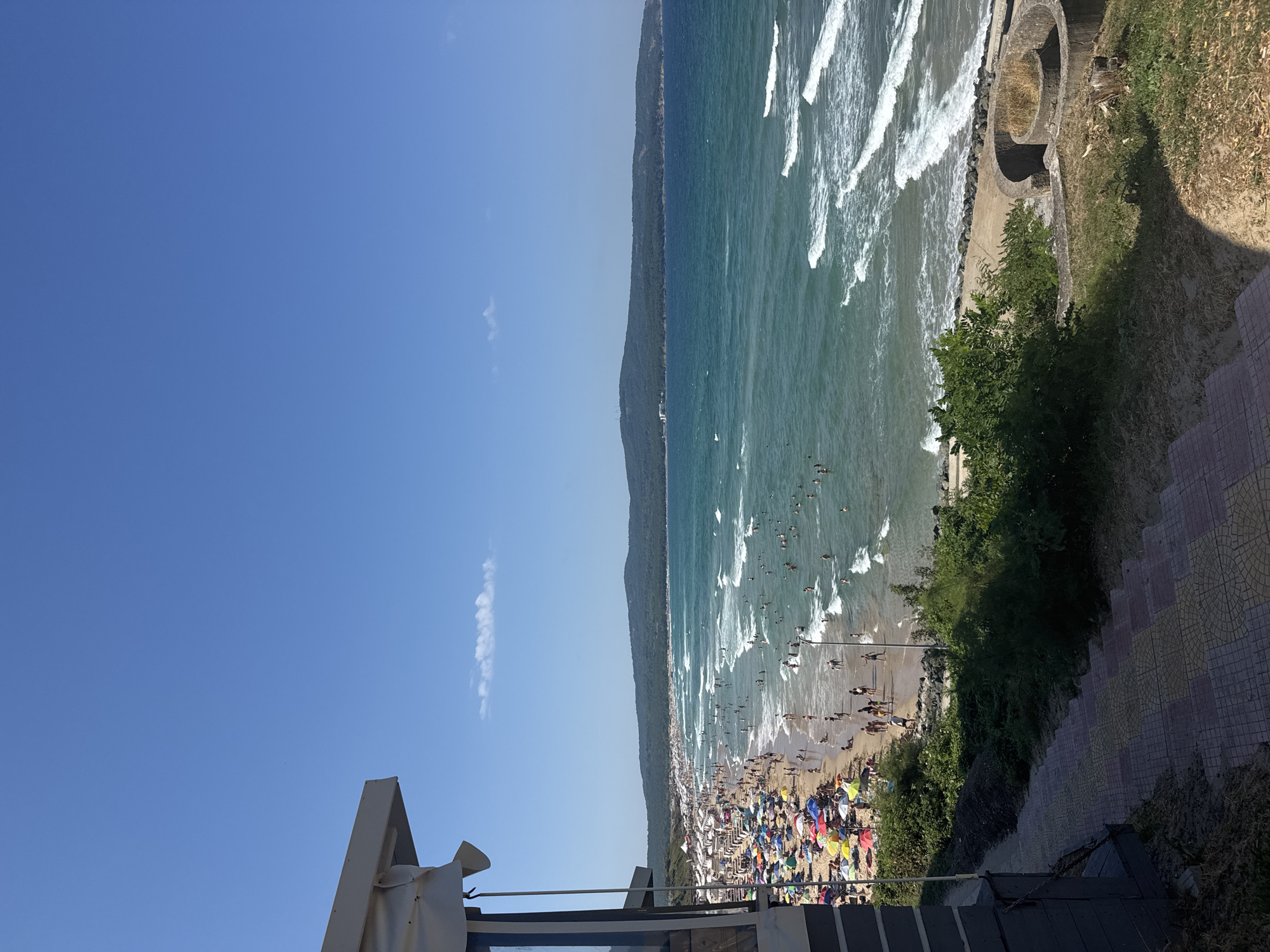 This is a high-angle, full shot of a crowded beach in Primorsko, Bulgaria, under a clear blue sky. In the foreground, a section of a modern building with a beige awning is visible on the left, and a tiled outdoor staircase leads down to the right. Beyond the stairs, lush green bushes and vegetation cover a slope leading towards the beach. The beach itself is densely populated with numerous people sunbathing, walking, and swimming in the turquoise sea. Colorful umbrellas are scattered across the sand, providing shade. Gentle waves with white foam break on the shore, and further out, the sea extends towards a distant, forested hill that forms the horizon. The overall scene depicts a lively summer day at a popular coastal resort.