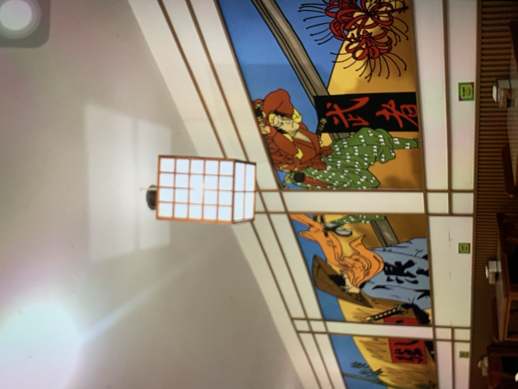 The image depicts a wall mural in what appears to be a restaurant, with the style of Japanese art. The mural features two samurai figures in action poses, with Japanese calligraphy prominently displayed. One samurai on the left wears a conical hat and a blue robe with dark characters on the back, holding a sword. The other samurai on the right wears a red top and a green patterned skirt, with a sword at his side. There are also depictions of red spider lilies and a yellow landscape. A modern, grid-like light fixture hangs from the ceiling. The lower portion of the image shows tables and chairs, suggesting a dining area. There is no identifiable information in the image that would place it in the city of Ciamis, Indonesia.