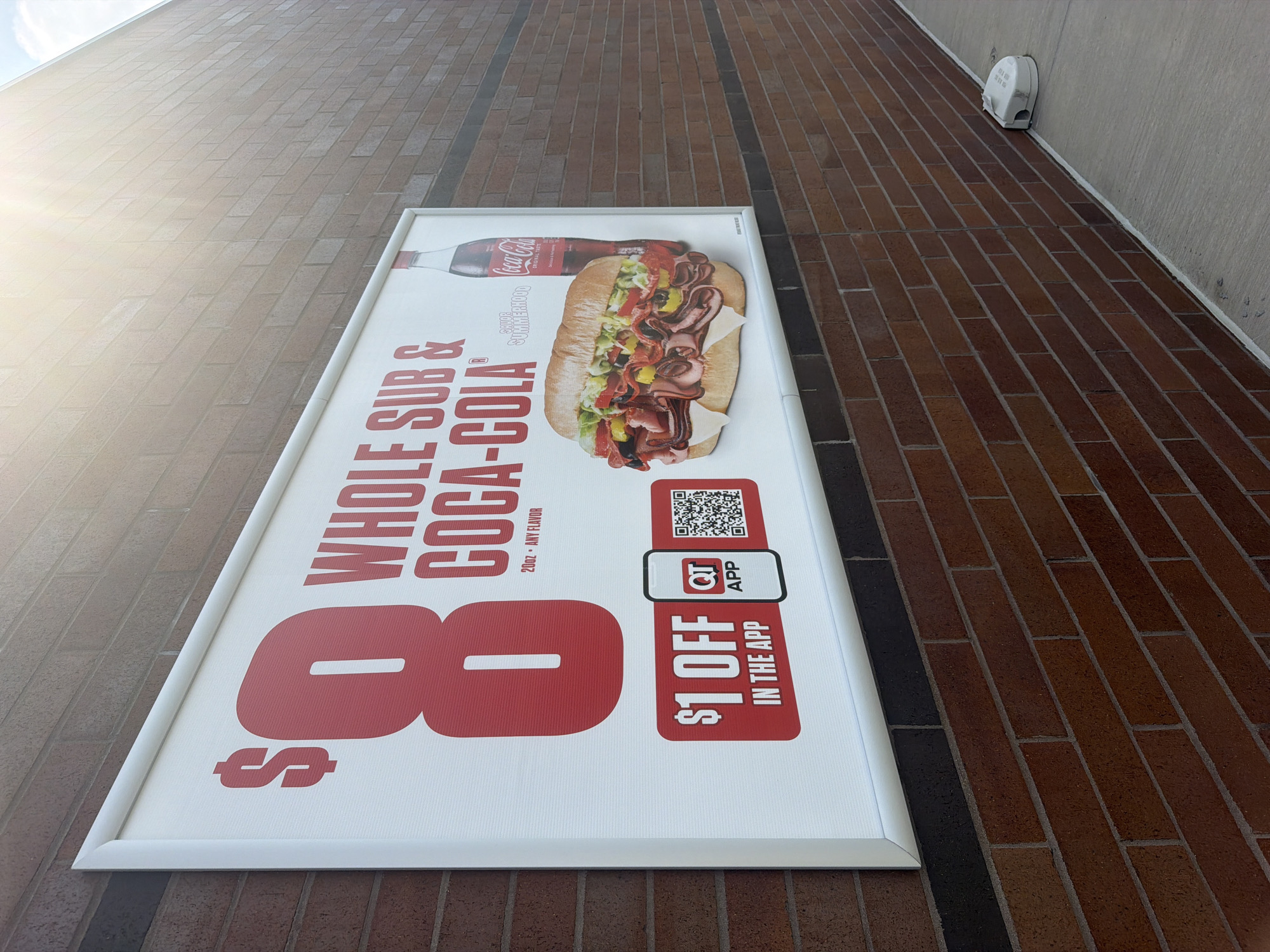 An outdoor advertisement is mounted on a brick wall. The advertisement is a large rectangular sign with a white background and red lettering. It promotes a "Whole Sub & Coca-Cola" deal for $8. A graphic of a large, filled sub sandwich is on the right side of the sign, next to a bottle of Coca-Cola. Below the deal information is a section that says "$1 OFF IN THE APP" with a QT app logo and a QR code. The sign is illuminated by sunlight from the upper right. The brick wall is made of reddish-brown bricks with dark mortar. The ground in front of the wall is a concrete sidewalk. A small, white robotic device is on the sidewalk to the right of the sign. The scene appears to be a commercial area.