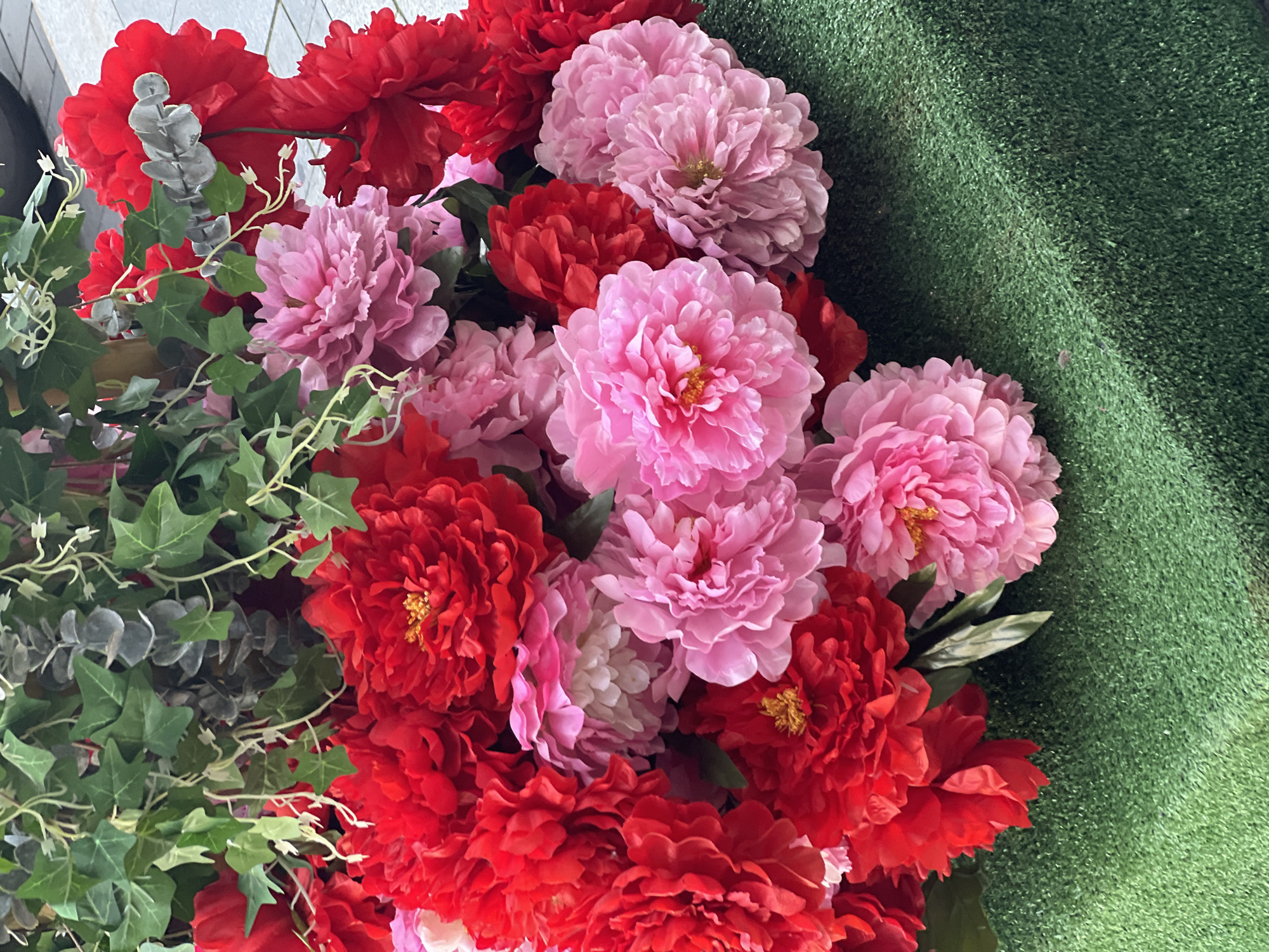 The image displays a vibrant, dense artificial floral arrangement composed primarily of large red and pink flowers, resembling peonies or dahlias, interspersed with various types of artificial green foliage. The flowers exhibit a range of colors, including vivid red and multiple shades of pink, from light pastels to brighter fuchsia tones. Many of the flowers feature prominent yellow centers.
The artificial foliage includes dark green ivy-like leaves with distinct lobes and lighter, grey-green rounded leaves reminiscent of eucalyptus, which are intertwined with and positioned above the flowers.
The lower section of the image shows a dark green artificial turf or grass mat, characterized by a textured surface that appears to be the immediate base or platform for the floral display. This turf exhibits slight variations in lighting and texture, suggesting a subtle contour.
In the upper background, particularly on the right side, a light grey paved surface is visible, indicative of an outdoor, urban setting. A dark, indistinct object, potentially a vehicle component or a decorative planter, is partially visible in the upper right corner.
No individuals are present in the scene, and no specific actions or interactions are depicted. The environment appears to be an urban outdoor space with a decorative floral installation. There are no specific visual indicators within the image to confirm the location as Moscow, Russia.