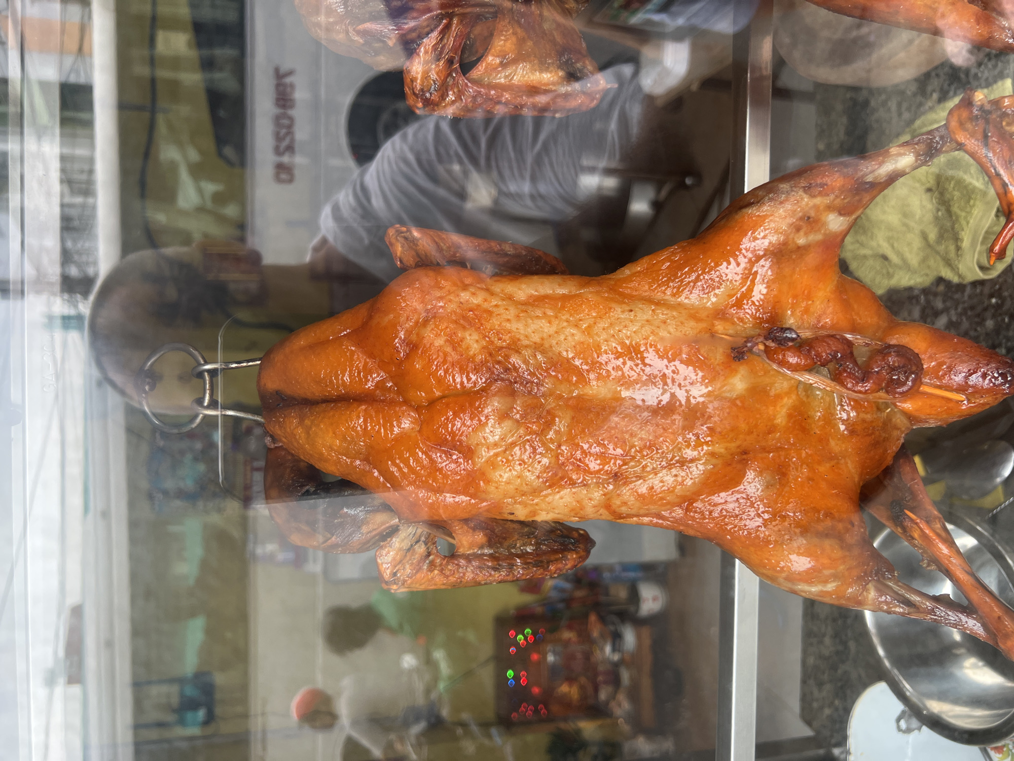 A whole roasted duck hangs in a glass display case, its skin a glossy, deep orange-brown. The duck is suspended by a metal hook through its neck. The foreground is dominated by the duck, with its legs splayed downwards. To the left, a stainless steel bowl is partially visible. In the background, through the glass, reflections show a street scene. A person with a bald head and wearing a grey shirt is reflected, appearing to be the establishment's worker. Other blurred figures and elements of a busy street, including a bus and signage, are visible in the reflection. This scene is located in Nha Trang, Vietnam, likely in a restaurant or market stall specializing in roasted meats, as indicated by the display of the duck. The overall impression is of a food establishment ready for business, with the roasted duck as the main attraction.