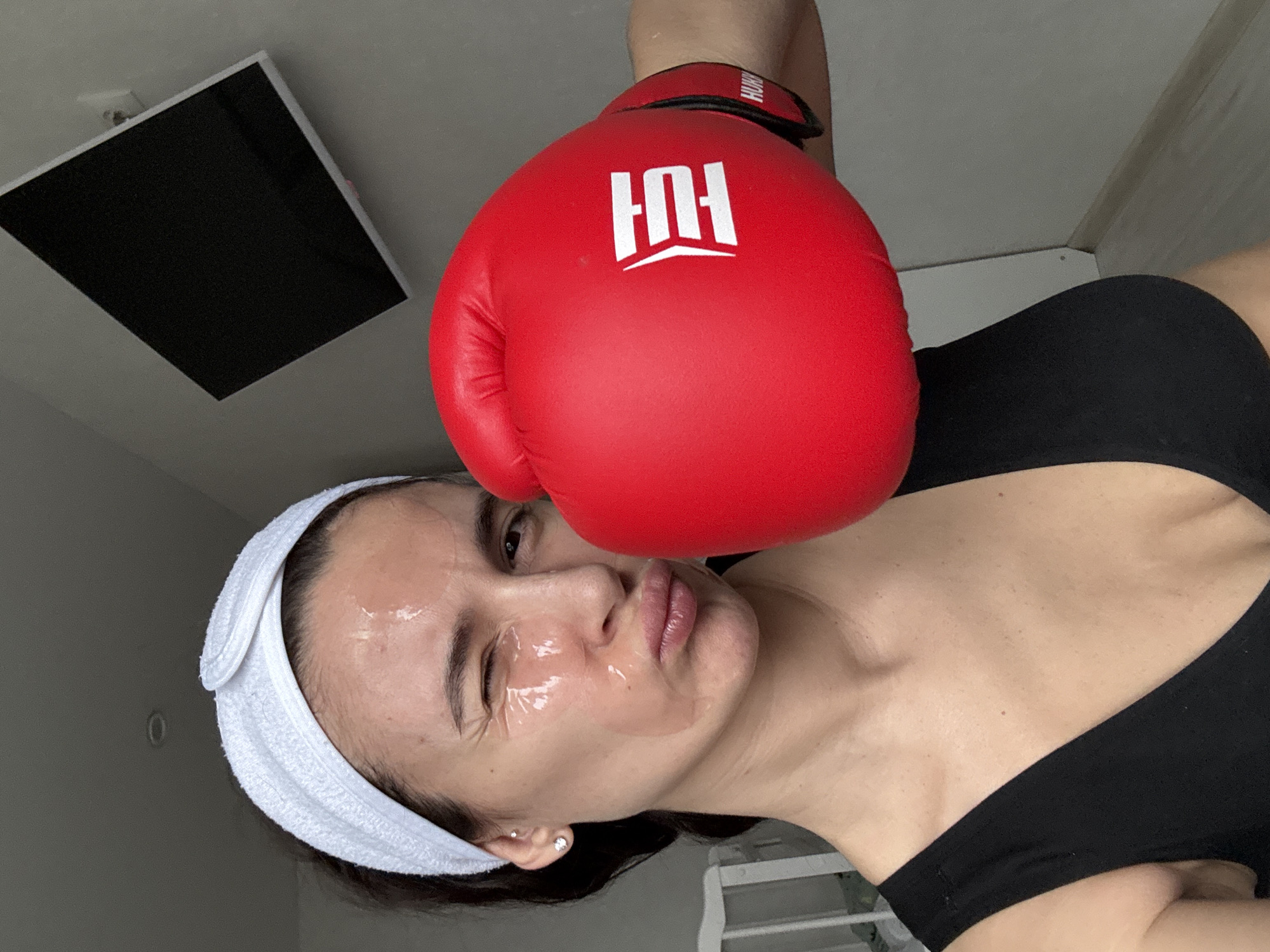 A woman is posing with a red boxing glove near her face. She has a white headband and a clear substance on her face, likely a face mask. She is wearing a black top. An open ceiling panel is visible in the background. The location is not specified.