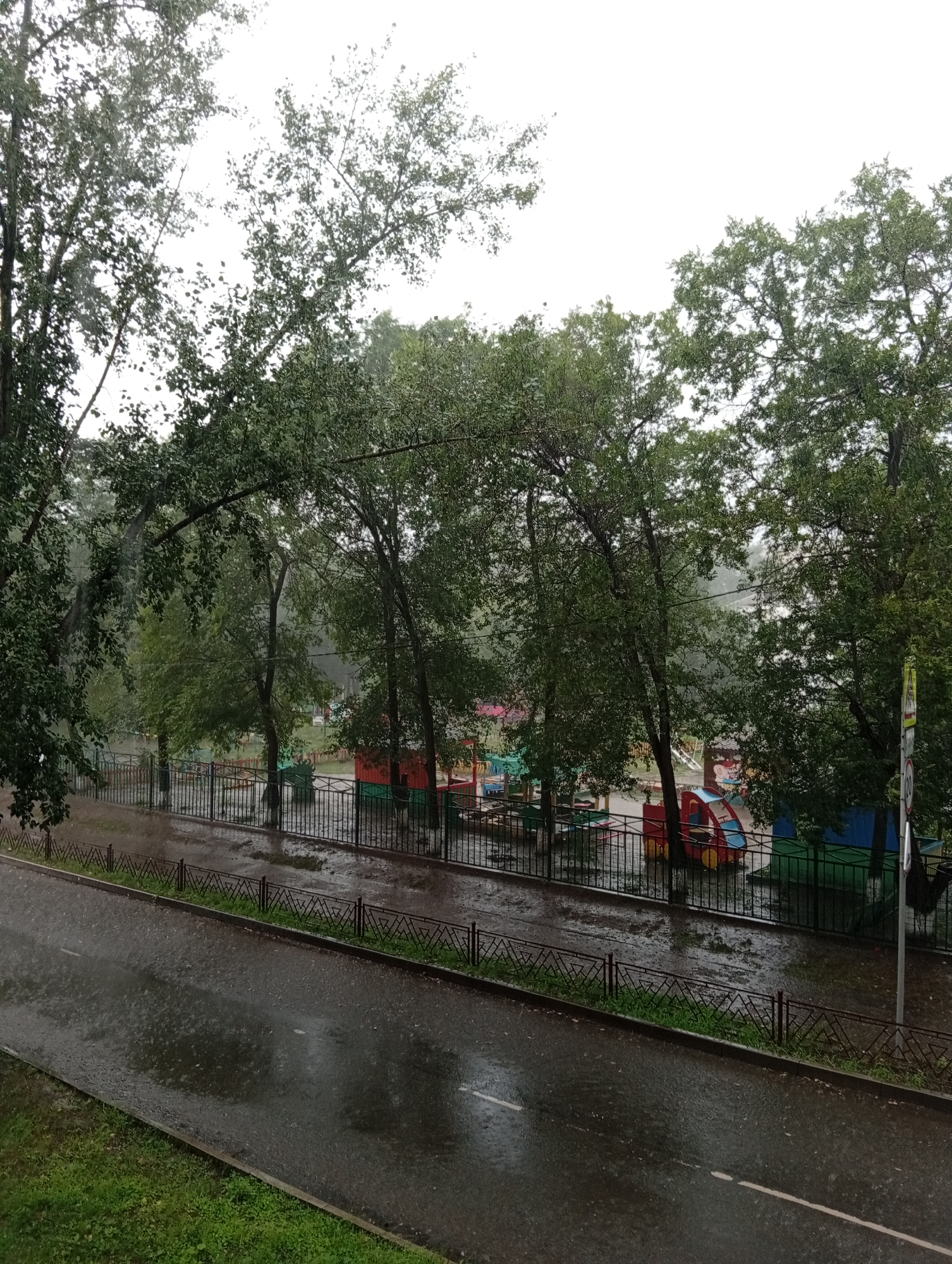 The image depicts a street scene in Minusinsk, Russia, during a heavy downpour. The sky is overcast, and rain is visibly falling, creating ripples on the wet asphalt road and sidewalk. Lush green trees with dense foliage line the street, their leaves appearing wet and dark. Behind the trees, a playground is visible, complete with colorful slides and swings. A black metal fence separates the playground area from the street. There are no people visible in the frame. The overall atmosphere is one of a stormy, wet day in a residential area of the city.