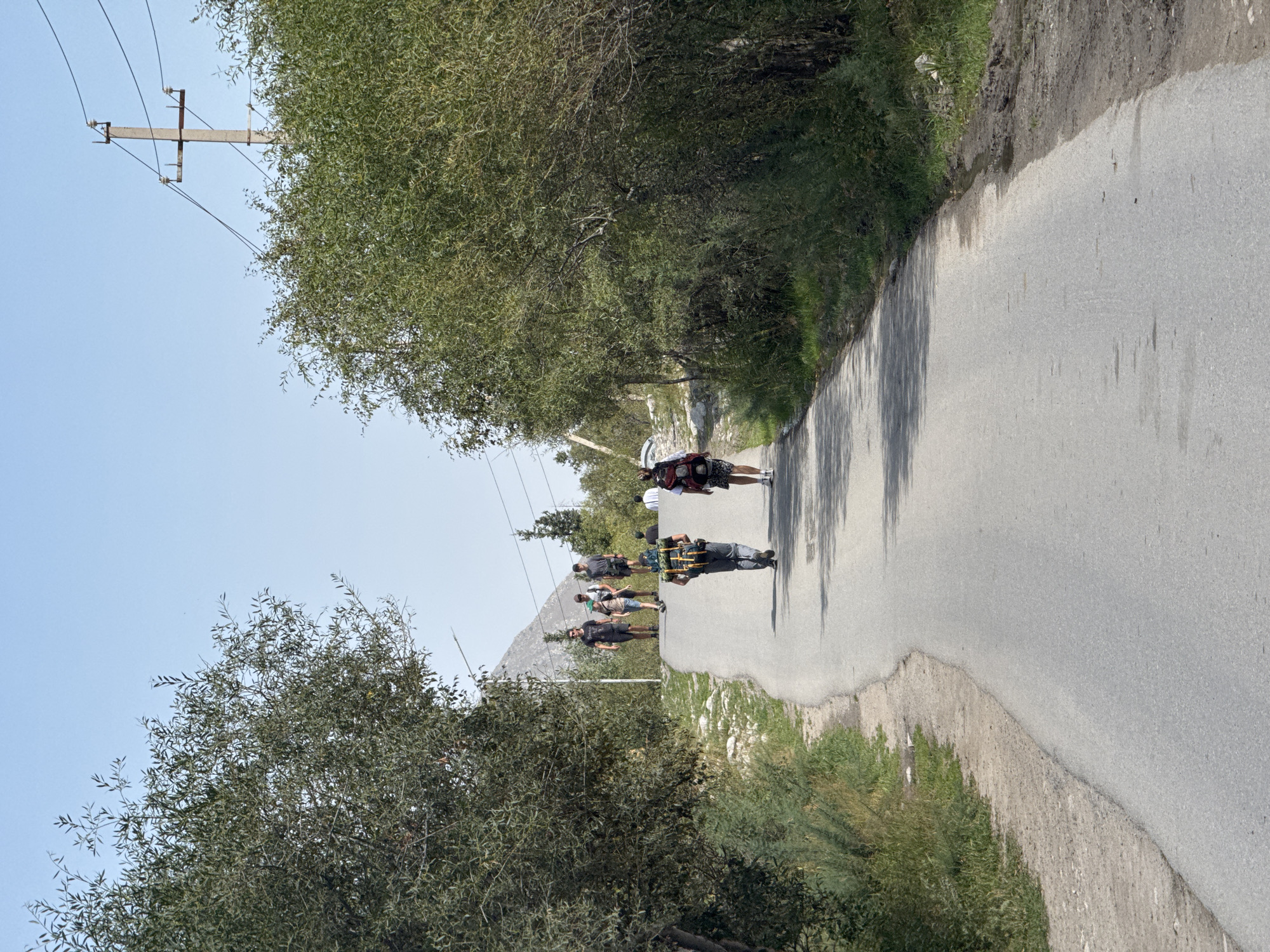 A group of hikers with backpacks walk up a paved road surrounded by trees and sparse vegetation. The road curves slightly upwards and towards the viewer's right. In the background, a clear blue sky is visible with a telephone pole and wires on the right side. To the left, a rocky, mountainous terrain is visible. The hikers are diverse in age and gender, all walking in the same direction. One hiker in the foreground wears a green and yellow backpack, while another in the middle ground wears a dark backpack with red accents. The trees lining the road are dense and leafy. The overall impression is that of a scenic hike in a rural or mountainous area of Kyrgyzstan.