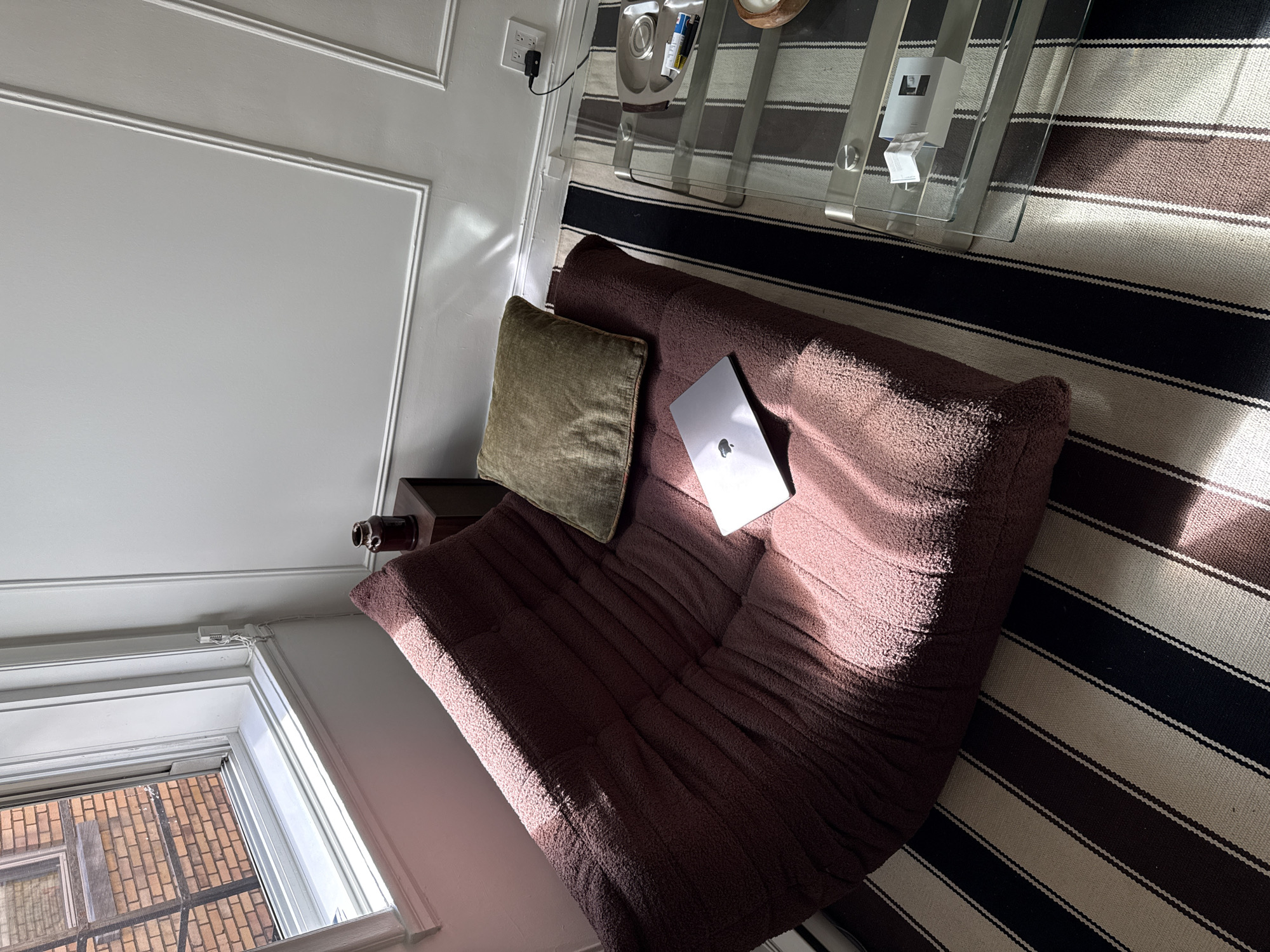 A high-angle shot captures a portion of a room interior, including a window that provides an external view of a brick building, likely indicating the location is in Montreal, Canada, given the architectural style. The window is positioned near the bottom-left corner of the frame, with an off-white wall above it. A cushioned brown sofa with a green pillow resting against the back occupies a significant portion of the central and lower areas of the image. A silver laptop is placed on the sofa seat. A dark-colored side table is situated to the left of the sofa. Adjacent to the sofa, a striped rug with dark and light horizontal bands covers the floor. In the upper-right corner, part of a glass-topped coffee table is visible, with various items on top. The wall on the upper-left side is painted white and features decorative molding. An electrical outlet and cord are visible near the top edge of the image, close to the coffee table. Sunlight casts shadows across the scene, particularly on the sofa and rug. No individuals are present in the image.