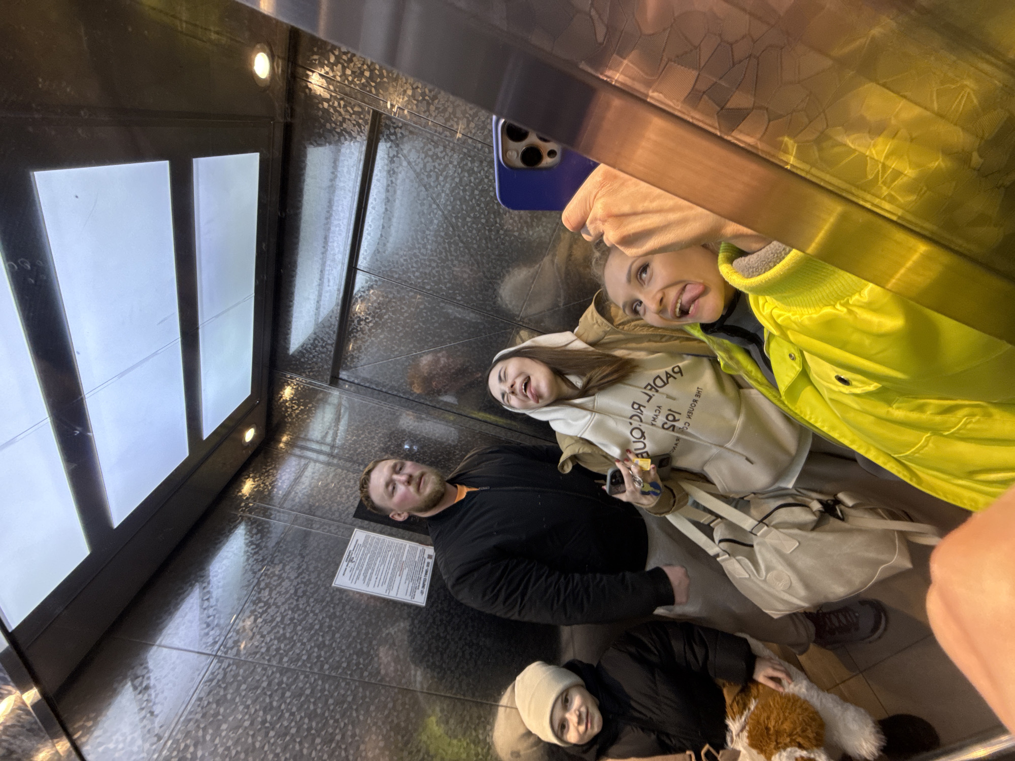 Four people are in an elevator, possibly in Perm, Russia. The elevator's interior is lined with patterned metal surfaces and has a row of rectangular lights. One person is taking a selfie with a blue phone. The person in the yellow jacket is sticking their tongue out. A man in a black jacket is positioned next to another woman in a white hoodie. At the bottom of the frame, another person is wearing a white beanie and a black jacket and holding a white and brown object, perhaps a dog.