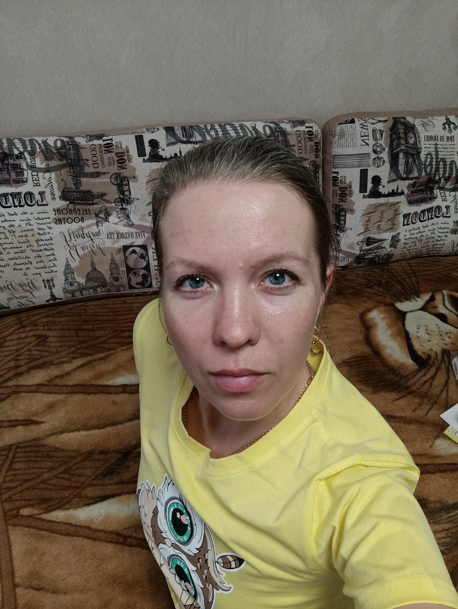 A woman with light skin and blue eyes is looking directly at the camera. She has fair hair pulled back from her face. She is wearing a yellow t-shirt with a cartoon owl design. A gold chain is around her neck, and gold hoop earrings are visible.
She appears to be sitting on a brown, patterned blanket, with a sofa behind her. The sofa has patterned cushions with images of London, including the Tower of London, the London Eye, Sherlock Holmes, double-decker buses, and other London landmarks. The patterned fabric appears to include Russian words in places. The image was taken indoors.