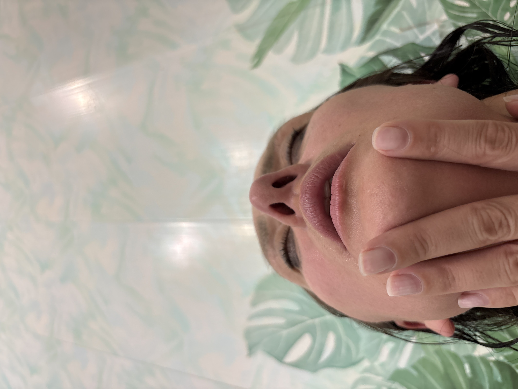 A close-up, rotated view shows a person, likely a woman, in a state of rest or sleep, possibly on a bed or sofa. Her head is positioned towards the right of the frame, with her face angled slightly upwards. Her eyes are closed, and her lips are slightly parted, revealing natural pink tones. Her right hand, featuring neatly manicured nails with a clear or light polish, gently covers the lower part of her face, from her chin to just below her mouth. Dark hair is visible on the right side of her head. The background is a soft, light-colored fabric patterned with large, light green tropical leaves, resembling Monstera leaves, suggesting an indoor, relaxed setting. The lighting is soft and even, highlighting the smooth texture of her skin. The overall scene conveys a sense of tranquility and relaxation, observed in Chaikovsky, Russia. No visible text is present.