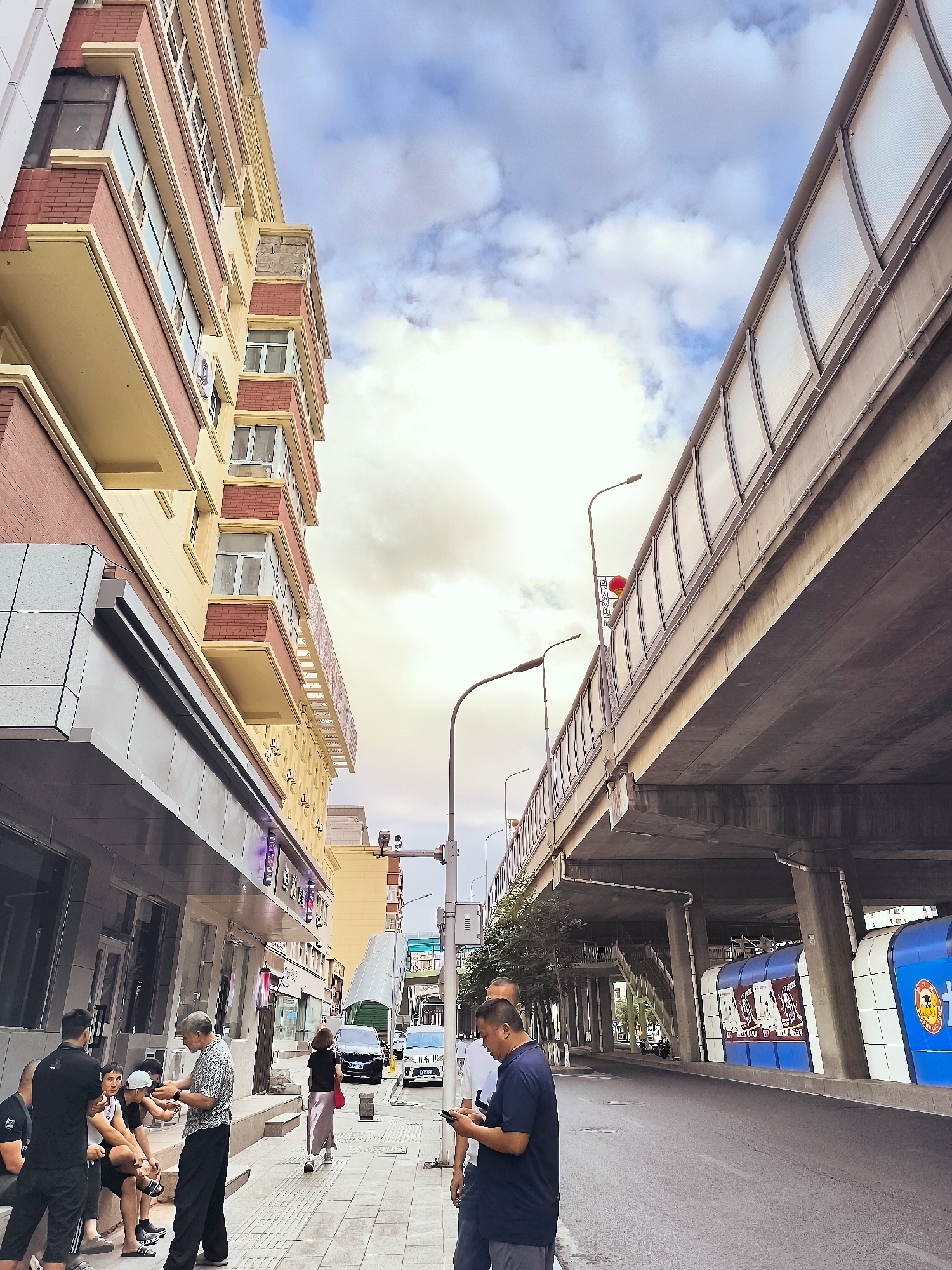 The image depicts a street scene in Ürümqi, China, under an elevated structure, possibly a highway or a pedestrian overpass. To the left, a multi-story building with red brick and yellow facade details lines the street. The ground floor features storefronts, with a barber shop visible. Several people are gathered on the sidewalk in front of the shops, some sitting and others standing.
The elevated structure dominates the upper portion of the image, with its concrete supports and underside visible. The top of the structure has a translucent barrier. Streetlights are present, including one with a red spherical ornament attached.
The street itself is paved, and cars are visible in the distance, including a black sedan and a white SUV. On the right side, beneath the elevated structure, there are large blue and white modular structures with posters featuring images of people.
The sky is partly cloudy, with patches of blue and diffused sunlight. The overall atmosphere suggests a typical urban environment.