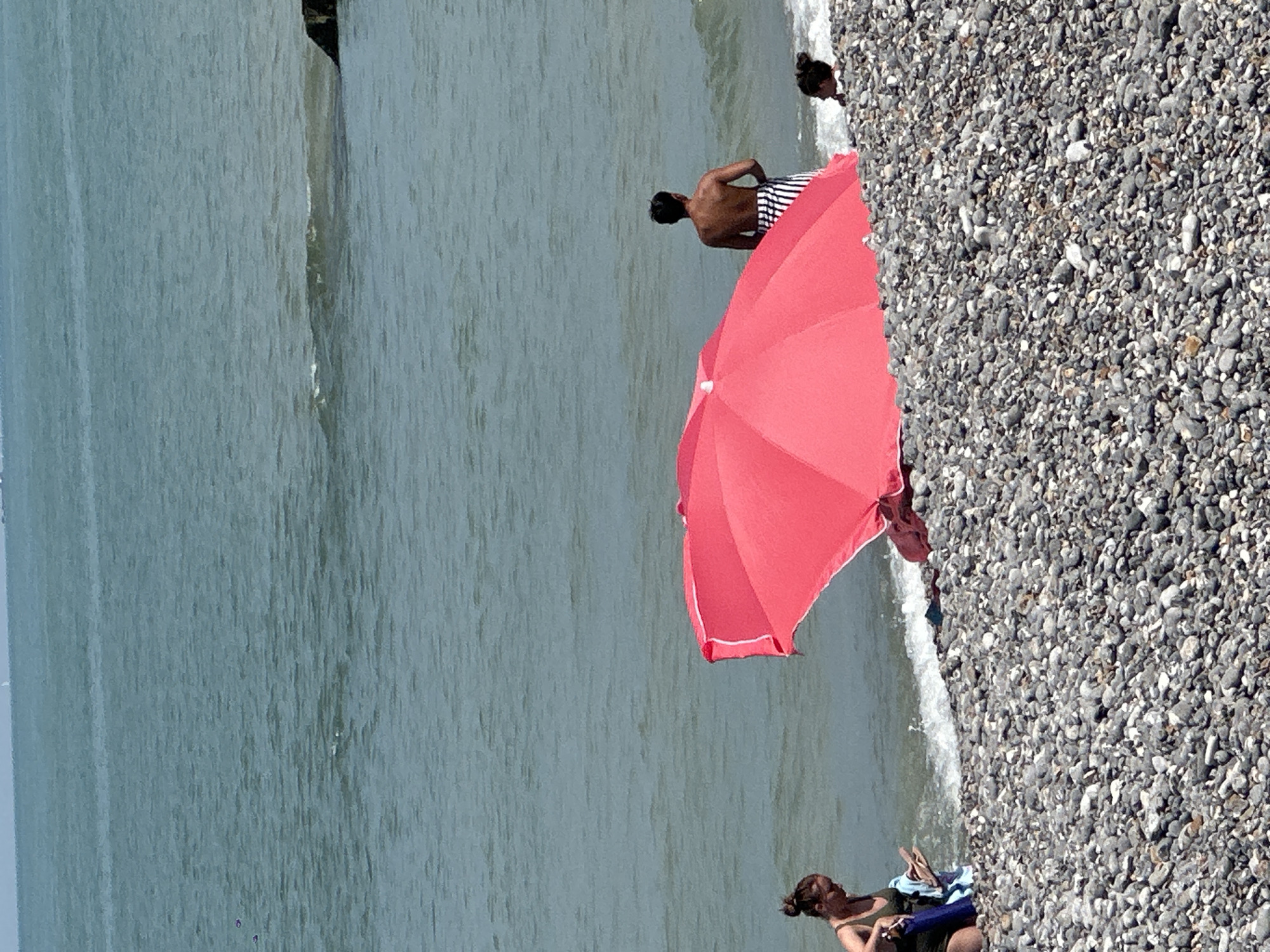 The image depicts a sunny day at the pebble beach of Hautot-sur-Mer, France. The water is a calm, blue-green, with gentle waves lapping at the shore. In the foreground, a vibrant pink beach umbrella is partially open, casting a shadow over the grey and white pebbles.
To the left, a woman with her hair in a bun is crouched down, wearing a dark green swimsuit and holding a blue towel and a pair of flip-flops. She appears to be getting ready to go into the water or has just come out.
Further out in the water, a man with dark hair is standing, facing away from the camera, wearing striped shorts. To his right, a child with dark hair pulled back is visible near the water's edge, their head just above the surface. The scene is populated by a few beachgoers enjoying the seaside.
