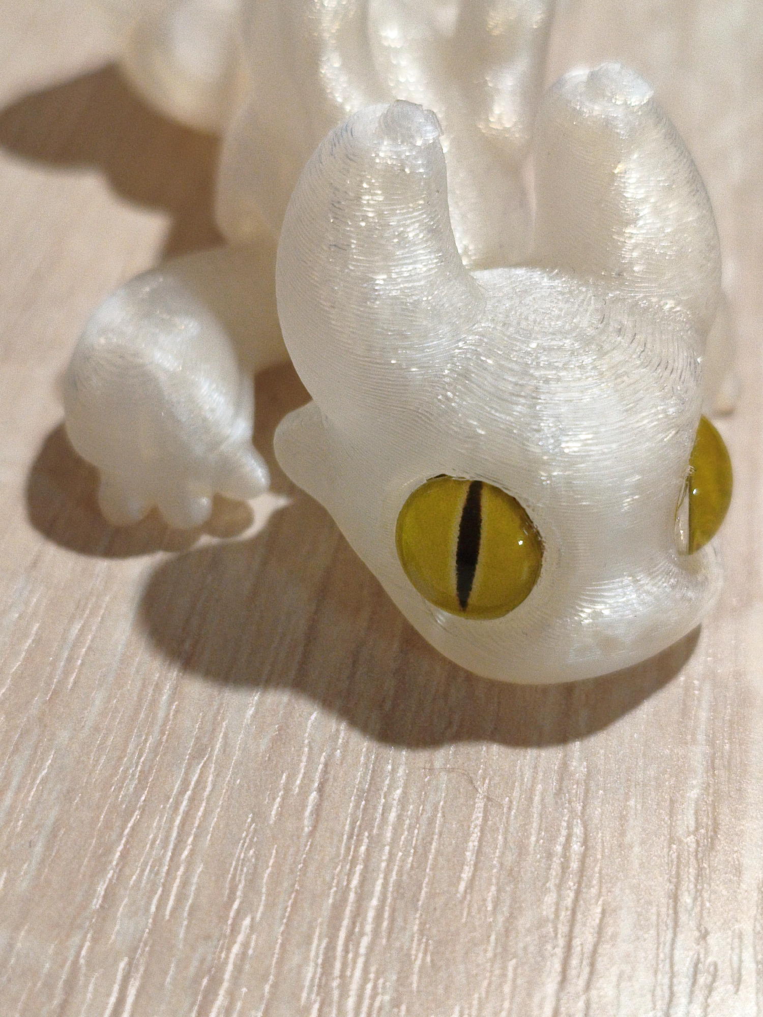 A close-up, slightly overhead shot shows a white 3D-printed figurine of a creature with yellow, slit pupils. The figurine is lying on its side on a light-colored, wood-grain textured surface. The shadow of the figurine is cast to the left. The material of the figurine shows visible layering from the 3D printing process, and it has a slightly translucent quality. There is no indication of location in Orenburg, Russia.