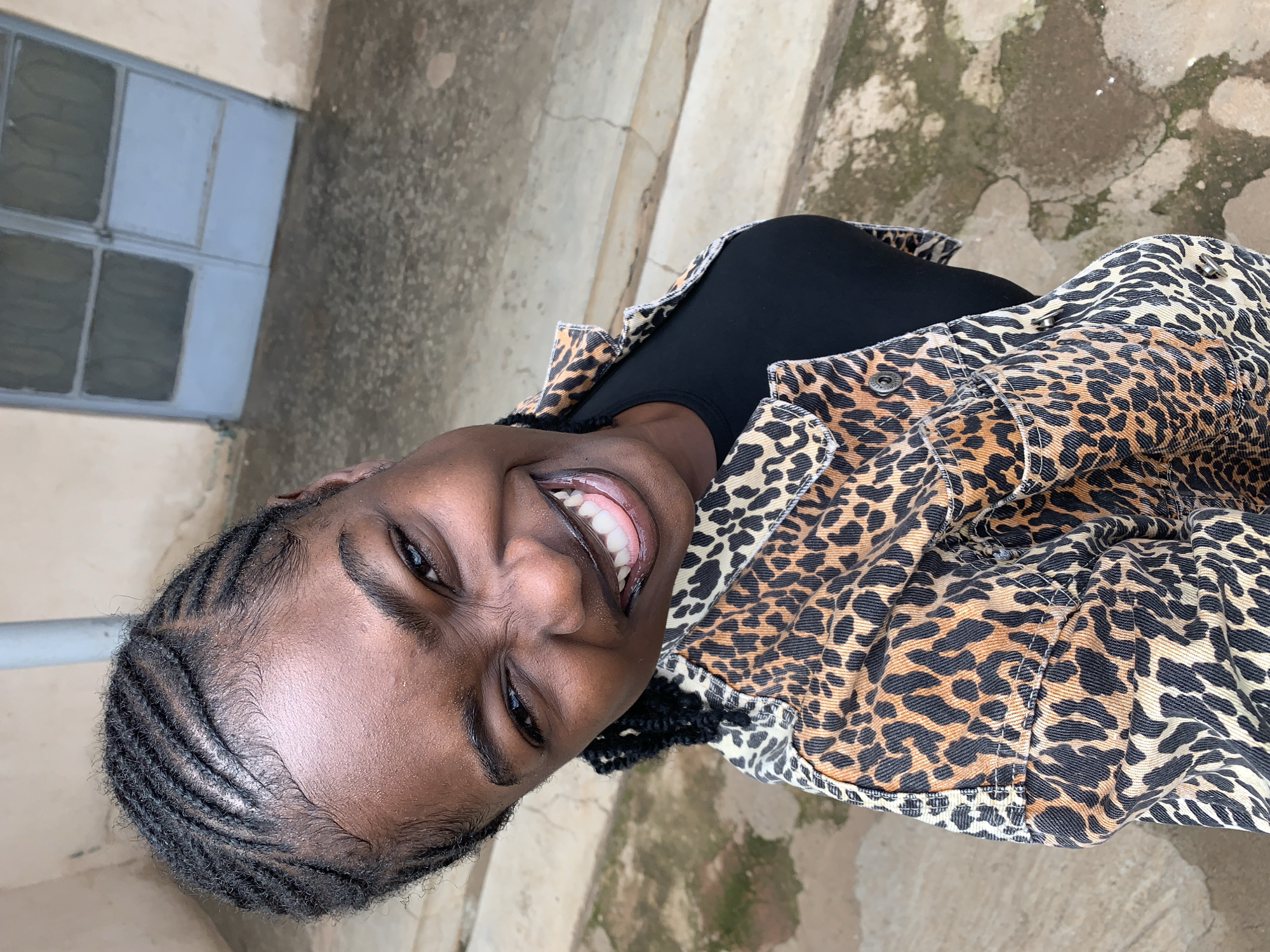 An overhead shot depicts a young Black woman smiling broadly and looking upwards directly into the camera. She has dark skin and her hair is styled in neat cornrows, with defined baby hairs along her forehead. Her expression is joyful, with crinkled eyes and a visible tongue between her white teeth. She wears a black top beneath a leopard-print denim jacket, featuring a collar, contrasting stitching, and metal snap buttons. The scene is outdoors in Rafin Par, Nigeria. Below her, the ground is an uneven, textured concrete or paved surface, exhibiting cracks and variations in color, suggesting wear and dampness. Behind her, a light tan-colored wall rises, with a weathered, light blue double-leaf door visible in the upper background. The door has two upper panels that appear to be mesh or frosted glass. A gray vertical pipe is positioned near the top left of the frame against the wall.