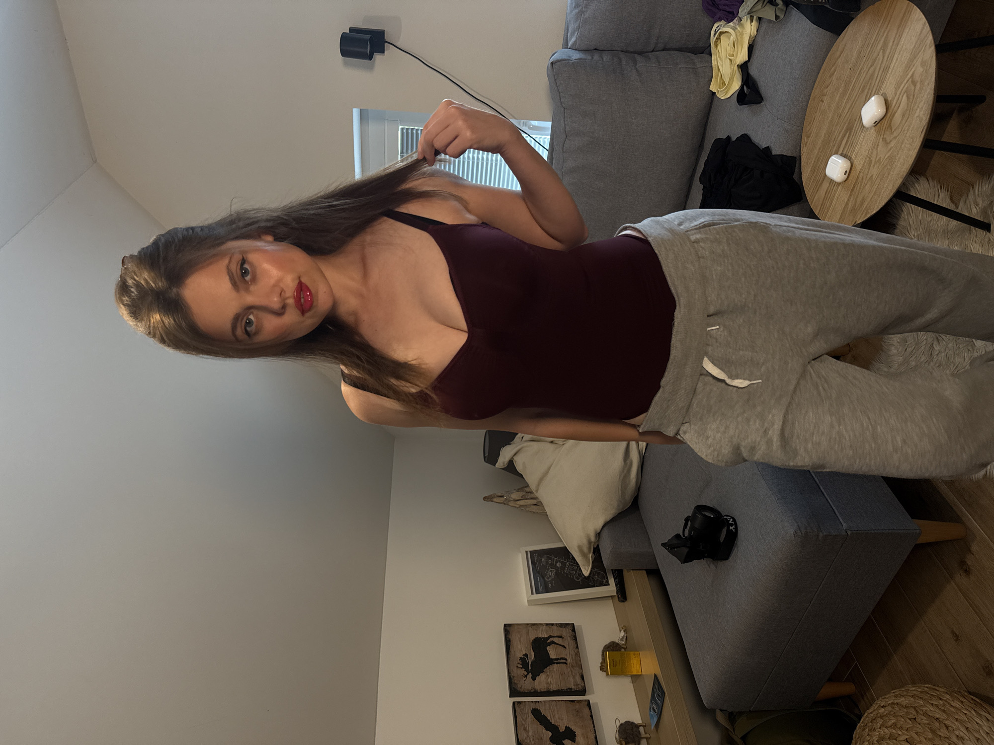 A woman stands in a room with slanted ceilings, suggesting an attic or loft space. She is wearing a burgundy camisole and grey sweatpants. Her hair is pulled up, and she has red lipstick on. To her left, a grey couch is visible, with a side table holding a camera. On the couch, a few items of clothing are scattered. A small, round wooden coffee table sits in the foreground with two white earbuds on it. The room appears to be a living space, likely in a residential setting. There is no indication of the specific city or country.