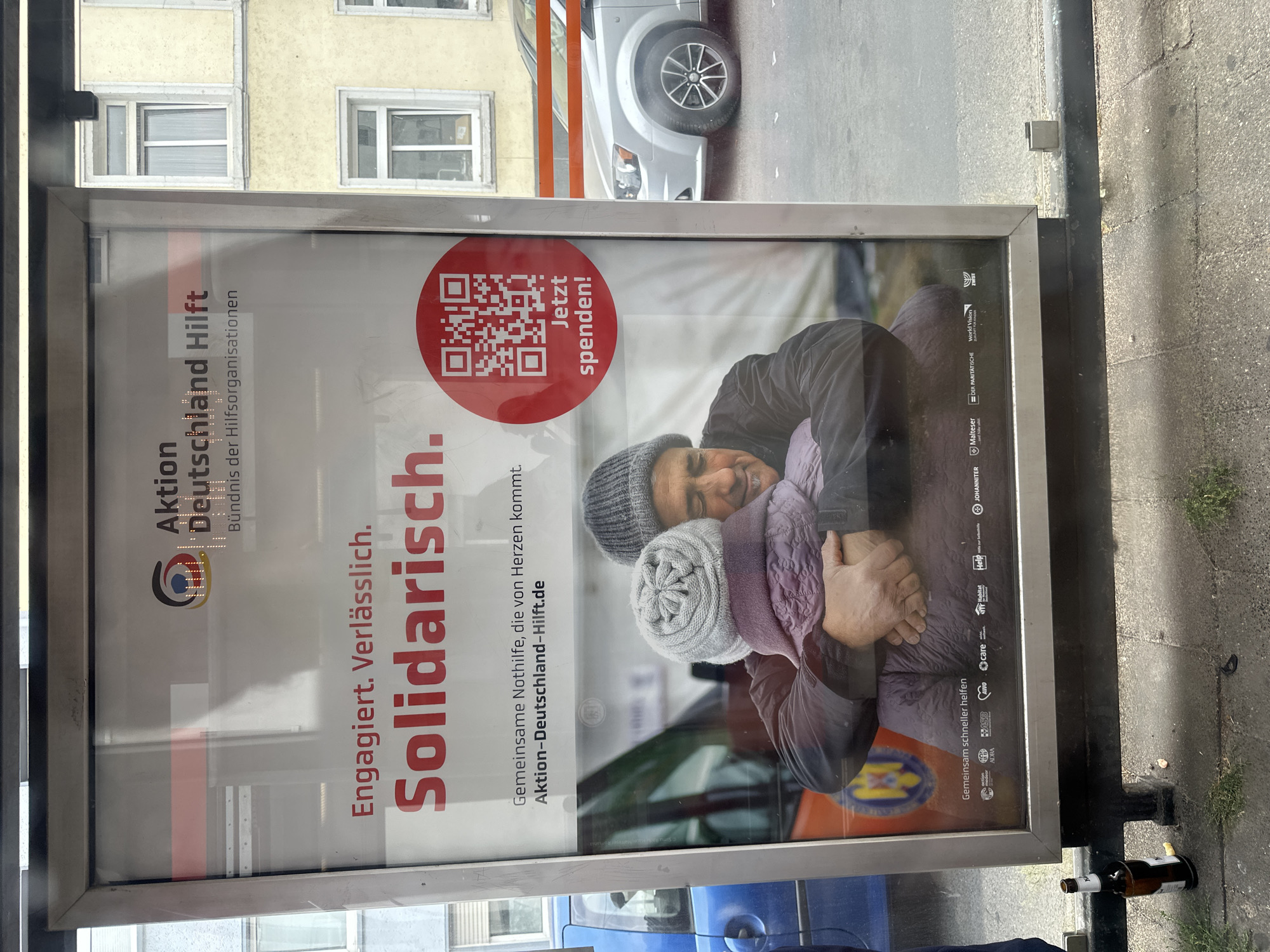 The image shows a poster on a bus stop shelter. The poster features a red QR code with the text "Jetzt spenden!" (Donate now!) next to it. Below the QR code is an image of two people embracing, appearing to be comforting each other. The poster has the heading "Aktion Deutschland Hilft" and the subheading "Bündnis der Hilfsorganisationen" (Alliance of aid organizations). It also contains the text "Engagiert. Verlässlich. Solidarisch." (Engaged. Reliable. Solidary.) and "Gemeinsame Nothilfe, die von Herzen kommt." (Joint emergency aid, which comes from the heart.) The website "Aktion-Deutschland-Hilft.de" is also visible.
In the background of the poster, there is a blue car on the left and a silver minivan on the right. The bus stop shelter has a metal frame and a glass front. Outside the shelter, on the ground, there is a brown beer bottle and some debris. The scene appears to be on a street with a sidewalk and road. The overall lighting suggests it is daytime.
While the poster itself promotes a national German organization, the specific location of the bus stop shelter within the city of Krefeld, Germany, cannot be determined from the visual information provided in the image. The buildings visible in the background are generic city architecture.
