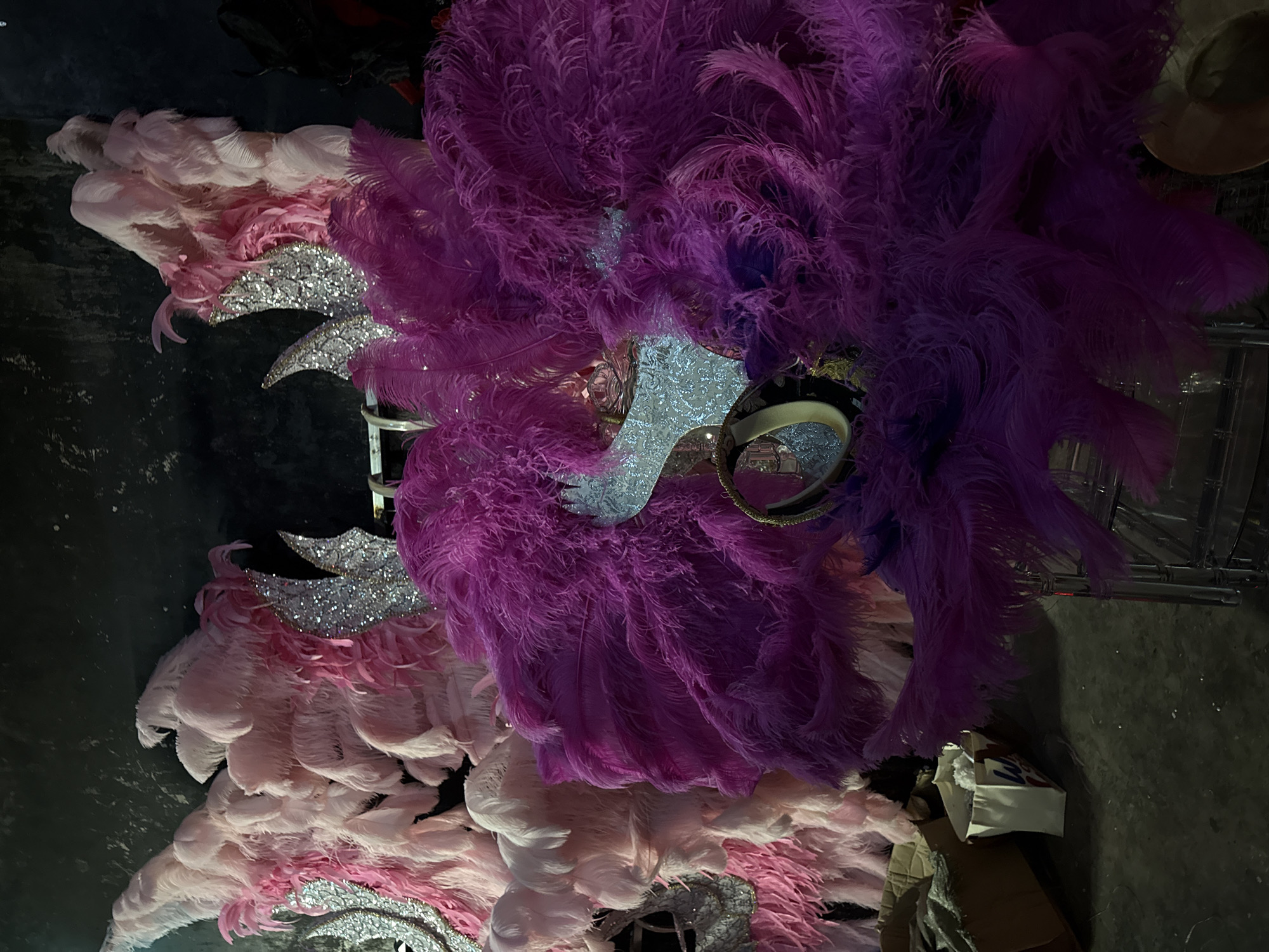 In Nanshan District, China, an indoor scene reveals an elaborate display of vibrant costumes, primarily composed of large, fluffy feathers in shades of pink and deep purple. Two prominent headdresses, likely part of a performance or parade, dominate the foreground. The one on the right is a large, circular arrangement of deep purple feathers, adorned with a shimmering, silver, horn-like structure and a circular gold band. To the left, a headdress made of lighter pink and white feathers is visible, also featuring silver, horn-like embellishments. The background is dark and appears to be a stage or storage area, with a textured dark wall. Cardboard boxes and a white paper bag are visible on the floor in the lower left of the frame.