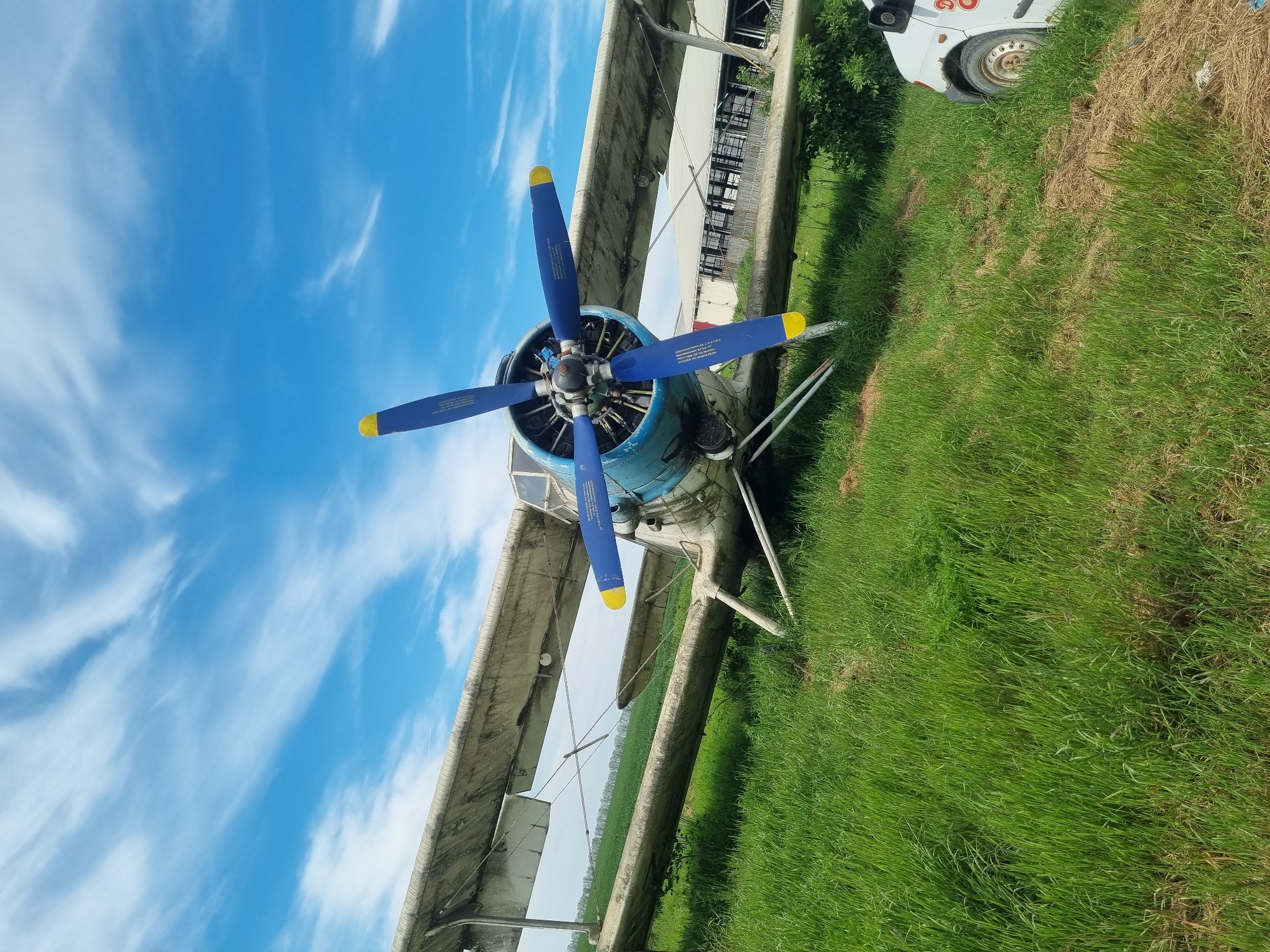 The image shows an Antonov An-2 biplane parked in a grassy field under a partly cloudy blue sky. The aircraft features a blue fuselage and blue propellers with yellow tips. The wings are painted in a faded gray or silver color. The background includes a building with a chain-link fence and the front end of a white van. The location is likely Ovcharovo, Bulgaria.