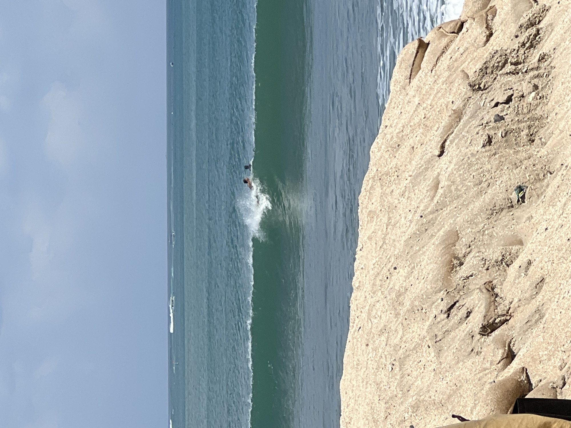The image depicts a scene at Kuta Beach, Indonesia, under a clear sky. The foreground is dominated by a sandy embankment, textured with small stones and shells. In the middle ground, a turquoise wave is breaking, with two individuals surfing on it. The surfer closer to the camera is in the process of riding the wave, with white spray around them, while the other individual is further back on the wave. In the background, the ocean stretches to the horizon, with a few distant boats visible.
