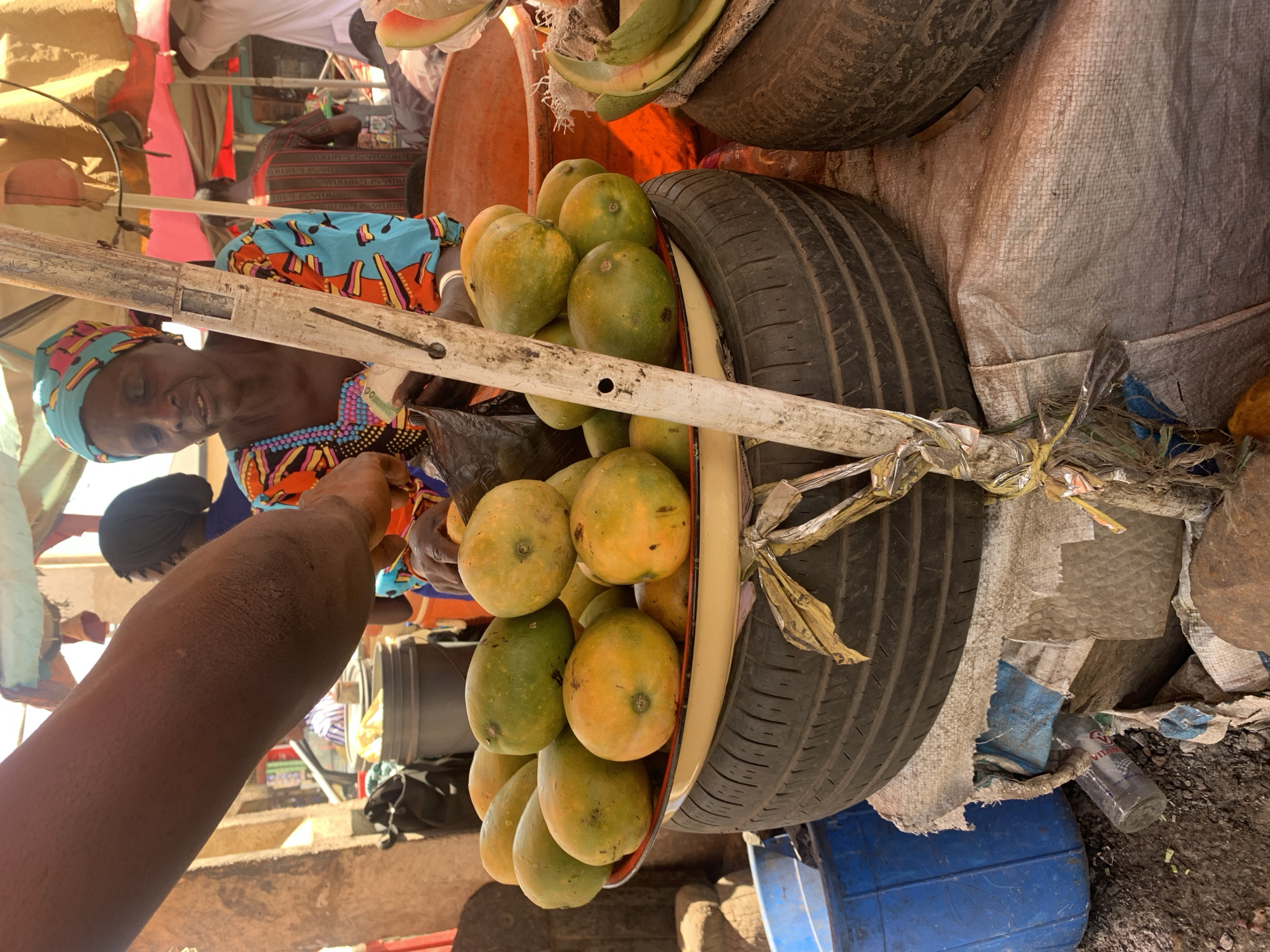 The image captures a market scene in Rafin Par, Nigeria. An elderly woman with a turquoise headwrap and colorful patterned top sits behind a fruit stand. A customer extends their arm towards her. The stand is constructed with a tire and a small bucket filled with mangoes. Another tire and what looks like some bananas are stacked above. A blue plastic drum sits below the stand.