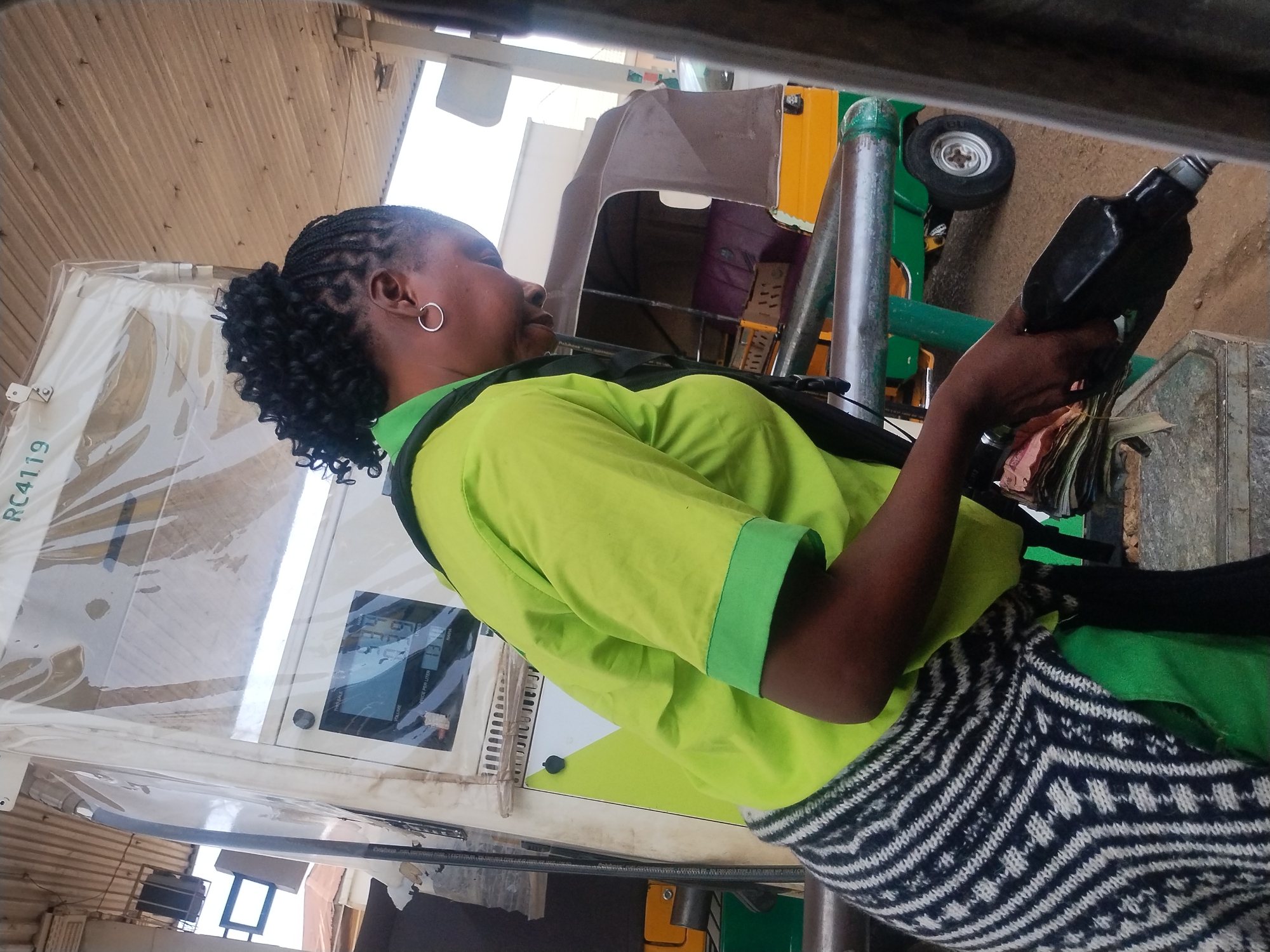A Black woman with dark braided hair in a bun and a hoop earring is standing at a gas station in Jos, Nigeria. She is wearing a lime green polo shirt with green trim on the sleeves and a black and white patterned skirt. She is holding a black gas pump nozzle in her right hand and a wad of cash in her left hand. Behind her is a gas pump with a digital display showing numbers. To the right, a yellow and brown auto rickshaw is visible. The scene is outdoors, with a corrugated metal roof overhead.