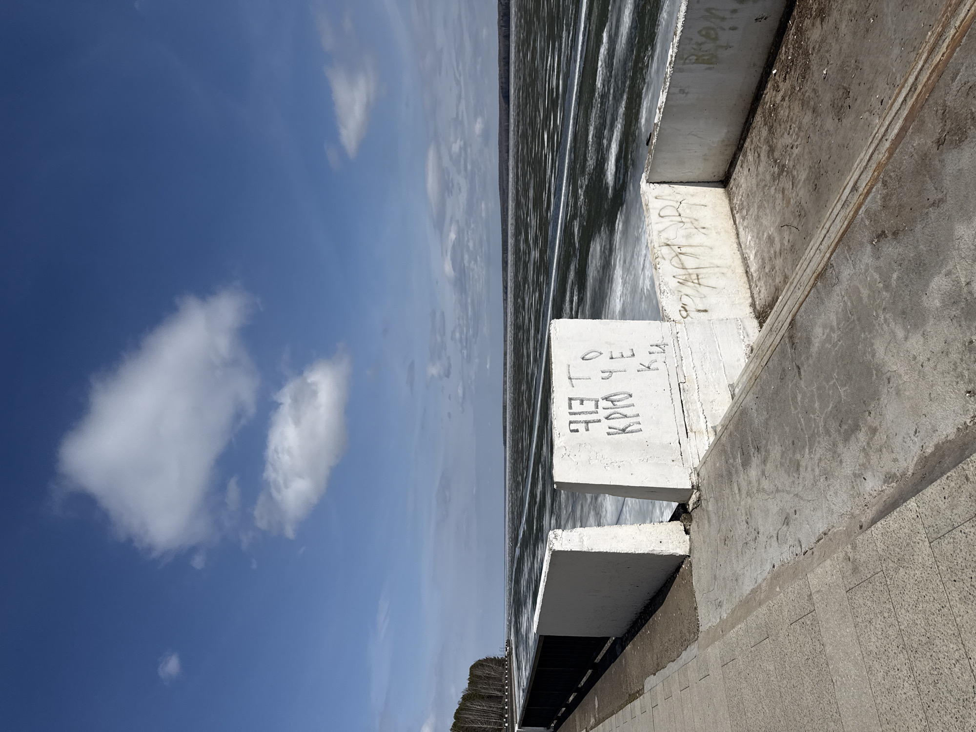 The image captures a daytime scene at a waterfront, likely in Chaikovsky, Russia. On the left, a bright blue sky is dotted with several white, fluffy clouds. The central part of the frame is dominated by a body of water, possibly a river or lake, displaying numerous small ripples across its surface, suggesting either wind activity or a gentle current.
To the right, a series of concrete structures forms an embankment or pier along the water's edge. The foreground features a large, light-colored concrete block, vertically inscribed with clear black Cyrillic letters: "ЭТО КРУТО" (ETO KRUTO), which translates to "THIS IS COOL". Behind this block, and extending further into the background, are more weathered concrete walls and slabs, exhibiting various textures and shades of grey, with some areas showing cracks and general wear. Faint, less decipherable graffiti is also visible on these rougher concrete surfaces.
In the far distance, beyond the water, a dark treeline stretches across the horizon, indicating a distant shore or landmass. The overall impression is one of bright, clear weather. There are no people visible, and no specific activity is taking place, presenting a calm, expansive view of a waterfront area.