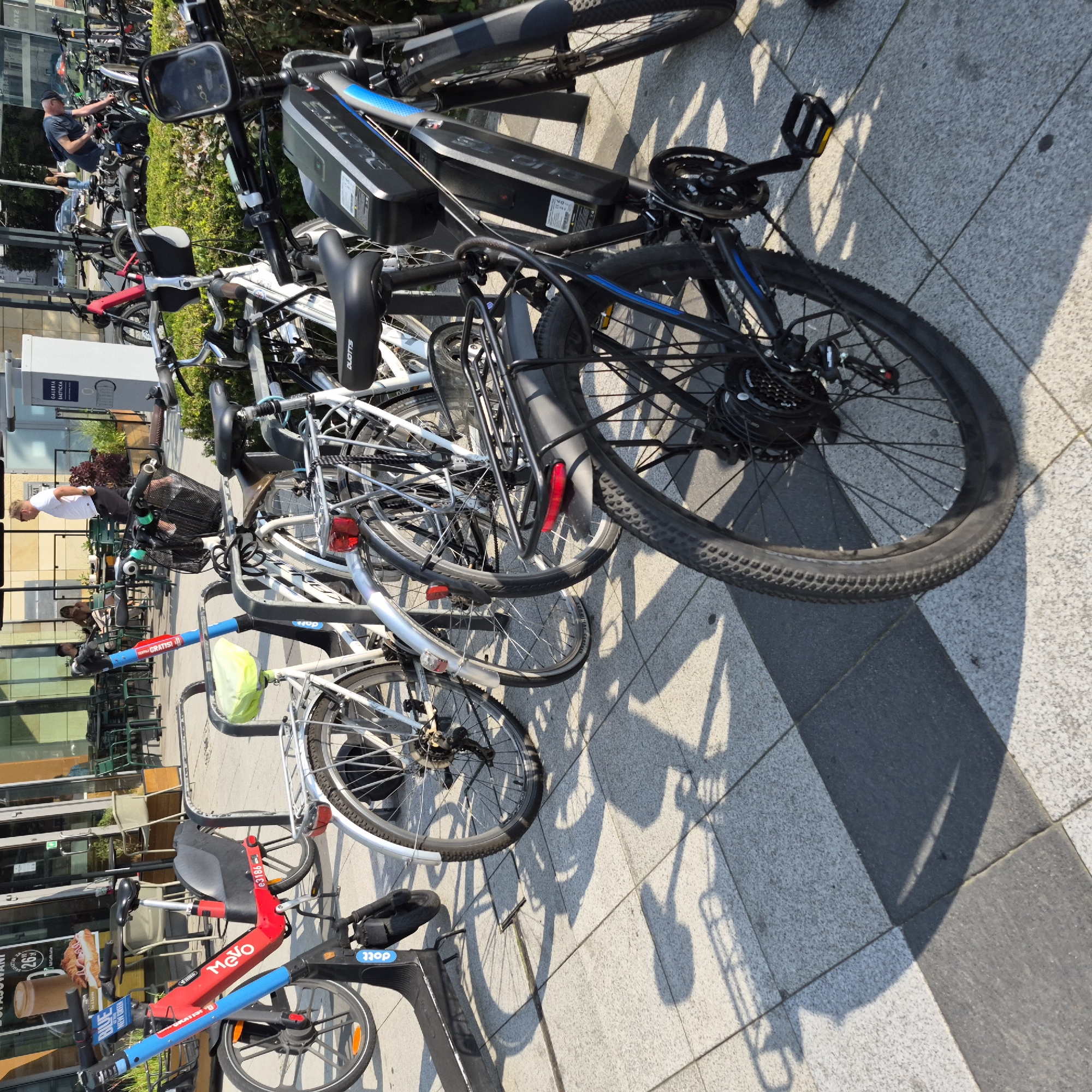 The image depicts an outdoor scene at Galeria Bałtycka in Gdańsk, Poland, likely near a cafe or restaurant area. A row of parked bicycles and an electric scooter is visible, suggesting a bike-sharing service. In the foreground, a black electric bicycle with a prominent battery pack is parked. To its left, a red and blue electric scooter with "Mevo" branding is also parked. Behind these, several other bicycles are lined up in racks.
Several individuals are present in the background. A man in a white shirt and dark pants stands near a table, looking towards the right. Another person, wearing a dark t-shirt and a cap, is seen bending over a bicycle, possibly inspecting it. Further in the background, more people are seated at tables, suggesting a public gathering or dining area.
The environment includes tiled pavement, planters with greenery, and the facade of a building with large glass windows. The lighting indicates it is daytime, with clear shadows cast on the pavement by the vehicles and people. The overall impression is of a functional urban space where people can access transportation and amenities.
