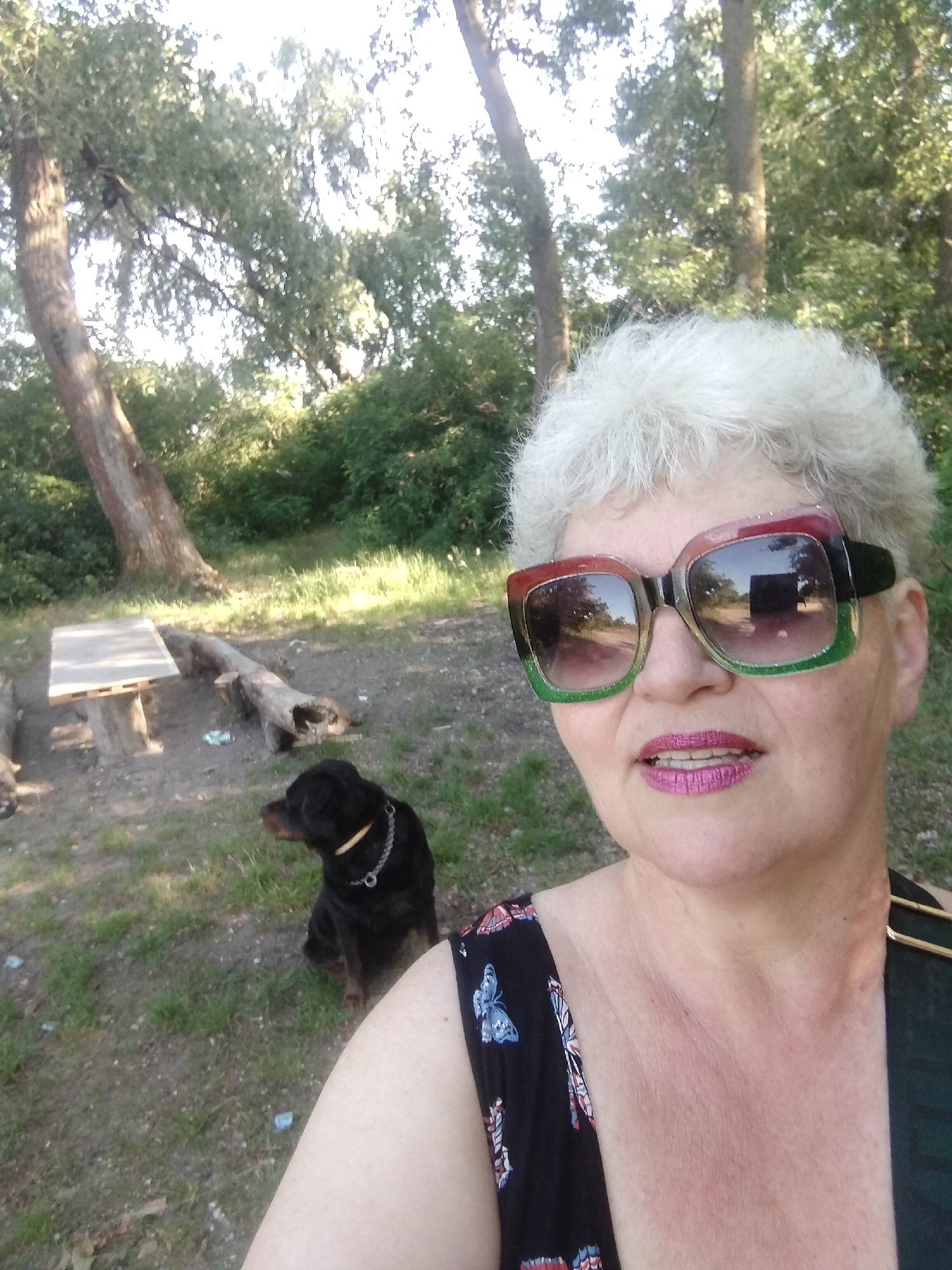 A woman with short, grey hair and large, colorful sunglasses is taking a selfie outdoors. She is wearing a black dress with a butterfly print. A black Rottweiler dog is sitting in the background to her left, facing away from the camera. In the middle ground, there is a wooden picnic table and a fallen log. The background consists of trees and greenery, suggesting a park or wooded area. The scene is likely located in Horishni Plavni, Ukraine, given the provided location context.