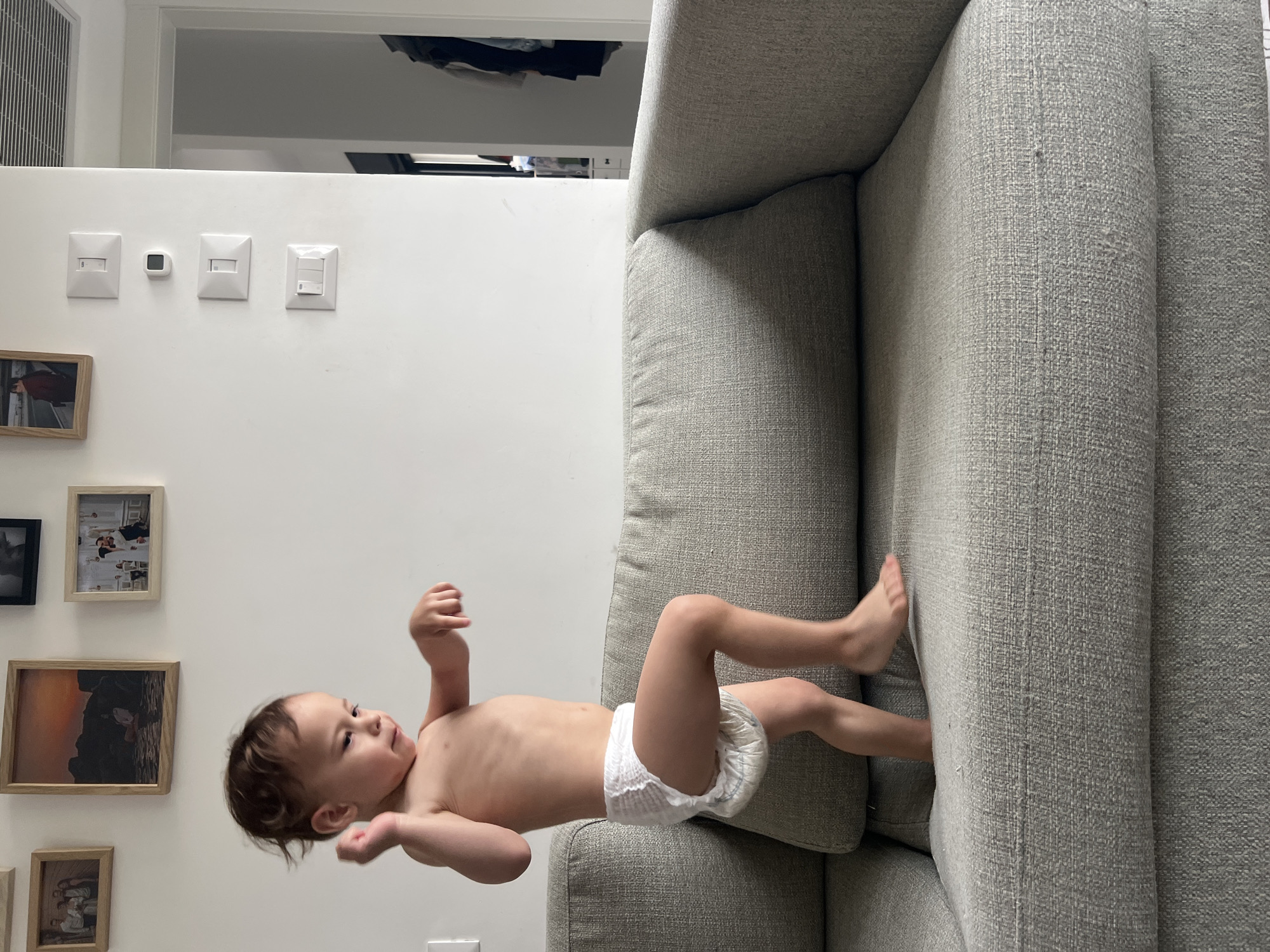 A baby wearing a diaper is on a gray sofa. The room has white walls with light switches and framed pictures. The location is Kiryat Bialik, Israel.
