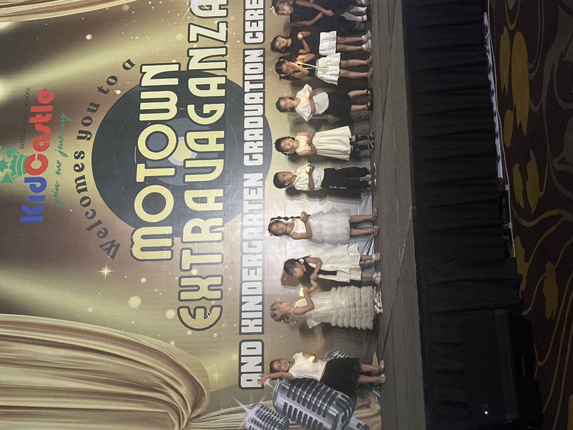 This media file captures a graduation ceremony for kindergarteners at KidCastle International School, as indicated by the text on a large banner. The banner, predominantly gold with sparkling accents, features the words "MOTOWN EXTRAVAGANZA AND KINDERGARTEN GRADUATION CERE," suggesting a theme that combines music and celebration. The scene is likely indoors, given the presence of a stage with black drapes and what appears to be carpeting with a distinct pattern.
The primary subjects are a group of young children, dressed in formal attire. Several girls are seen wearing white dresses, while some are in black and white outfits, holding small lights that resemble candles. Their expressions, though not entirely clear due to the image's orientation and resolution, seem to reflect the seriousness and excitement of the occasion. One child, in the foreground, is leaning back and looking towards the camera with a smile. Two large, vintage-style microphones are also visible at the bottom of the image, reinforcing the "Motown Extravaganza" theme and suggesting a performance or presentation aspect to the graduation. The lighting appears to be artificial, illuminating the stage and the banner. The location is specified as Nha Trang, Vietnam.