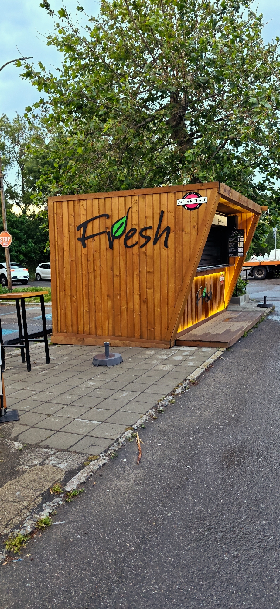 The image shows a "Fresh" stand located in Burgas, Bulgaria. The stand is constructed of wood and has the word "Fresh" written on the side, with the "e" replaced by a green leaf. The stand also has a "Cafés Richard" logo on the front. The stand is situated on a paved area next to a street. There is a table and chairs next to the stand. A stop sign is visible in the background. Cars and a truck are also visible in the background. The sky is cloudy.