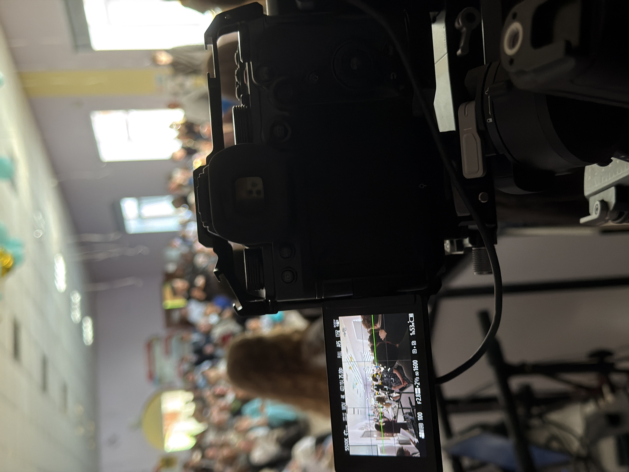 A digital camera on a rig is positioned to film a room filled with people. The room has white walls and a drop ceiling with fluorescent lights. The camera display shows a recording in progress. The recording setting are 5500K, NOV 4K 4770/114 25.00P FULL AFS E52. The recording length is 1.53m. The photo was taken at 100 ISO, f2.8, -2/3, so 1600. The location is unknown and the prompt did not specify it must be in the city of Krasnoyarsk, Russia.