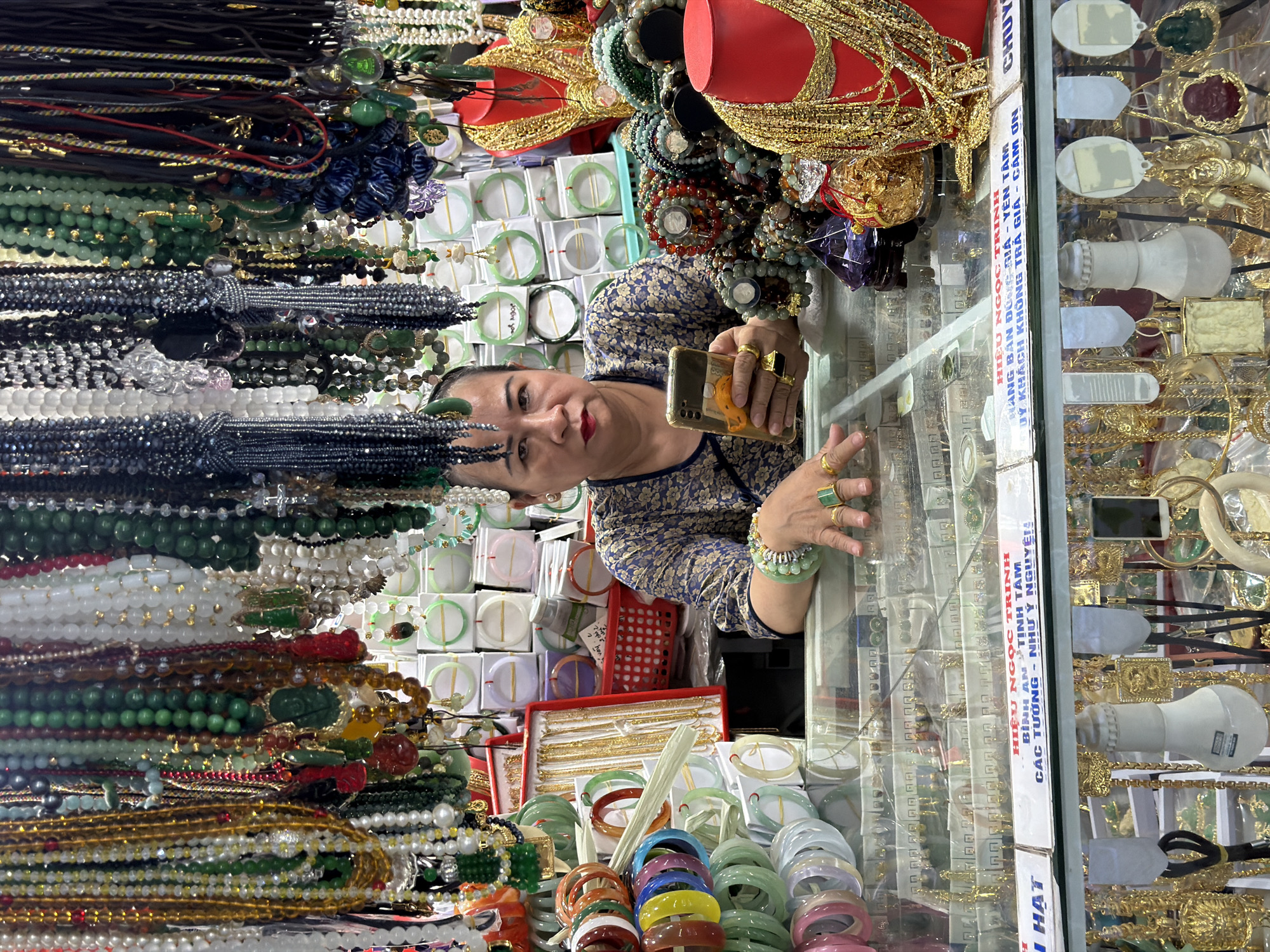 A woman is standing behind a glass display case full of jewelry in Đà Nẵng, Vietnam. She is holding a smartphone in her right hand and looking at the camera with a slight smile. She has several gold rings on her fingers and a green jade bracelet on her left wrist. The display case contains a variety of necklaces, bracelets, and rings, including gold, jade, and other materials. Many jewelry pieces are stacked on the left, while a row of jade bracelets lines the bottom center. Behind her and in the display case are rows of jewelry, small jewelry boxes, and other display items. The text "HIỆU NGỌC TRINH," "BÌNH AN TỊNH TÂM," and "CÁC TƯỜNG - NHƯ Ý NGUYỆN" are on the vertical divider of the display case.