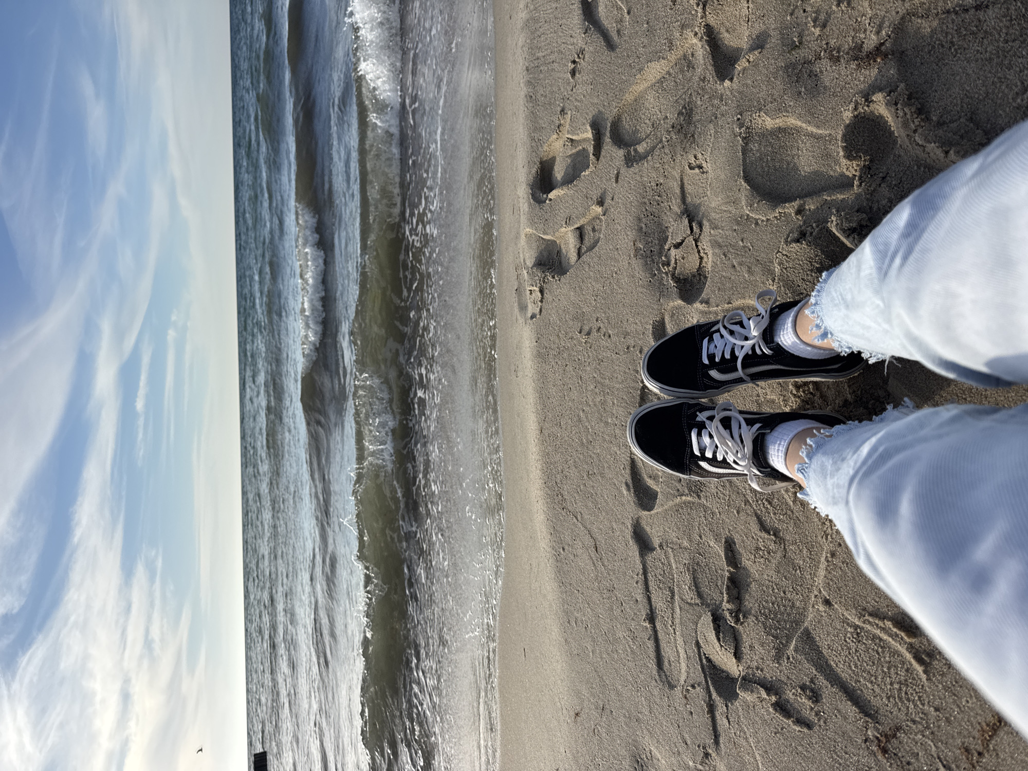 A person stands on a sandy beach with the ocean to their left. The person wears light-wash jeans, white socks, and black and white Vans sneakers. The sand is marked with footprints. The ocean is choppy with visible waves. Above the ocean, the sky is visible with scattered clouds. The image is rotated counterclockwise. The location is Priboy, Russia.