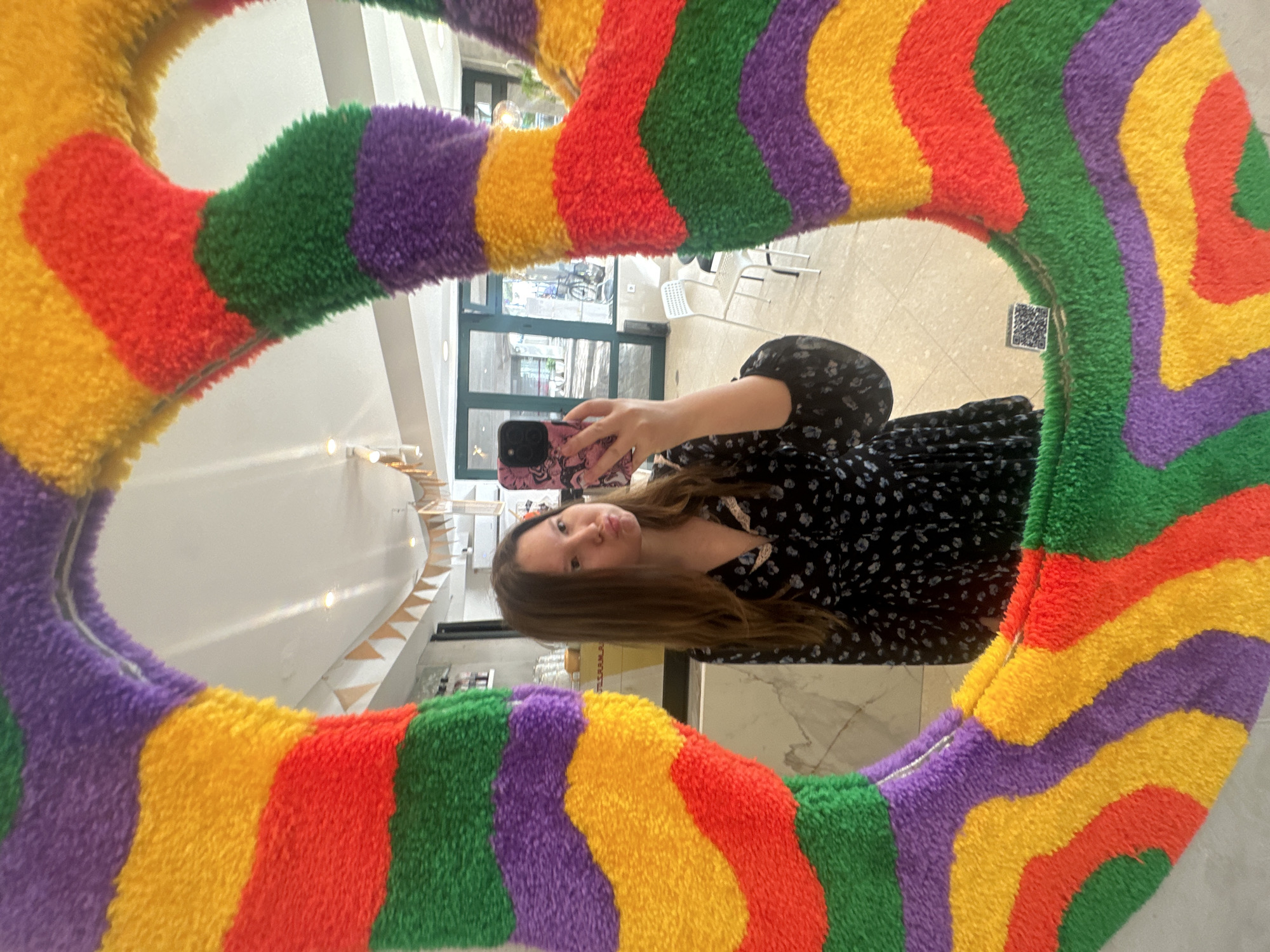 A young woman is captured in a mirror selfie, reflected at eye-level within a large, circular mirror. The mirror's frame is highly decorative and plush, featuring wide, wavy stripes in vivid colors: red, orange-yellow, green, and purple, arranged alternately. The woman has long brown hair and is wearing a dark dress with a subtle pattern of light-colored dots or flowers. She holds a smartphone with a dark case and a distinctive pink and red swirled design, featuring multiple camera lenses. Her lips are slightly pursed in a playful pout.
The mirror reflects an interior space, likely a modern shop or cafe in Budva, Montenegro. Through large glass doors or windows visible in the reflection, an outdoor area with white, modern-style tables and chairs can be seen, along with reflections of trees or nearby buildings. Inside, a light-colored wall features a wooden staircase or tiered display, with bright ambient lighting suggesting it is daytime. A small, black and white QR code sticker is adhered to a surface within the reflected background, positioned just inside the mirror frame to the right.