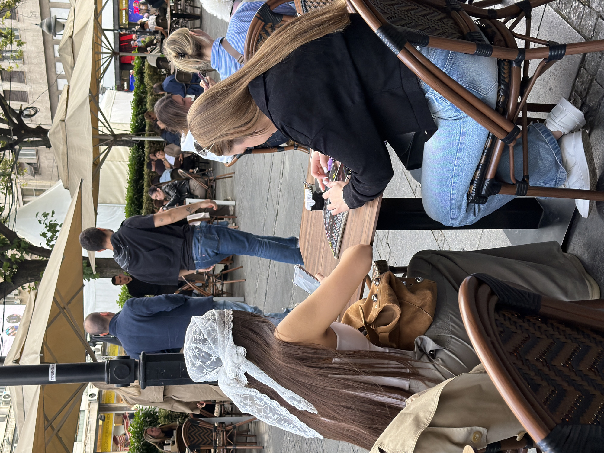 A medium shot captures a scene at a cafe, possibly in Belgrade, Serbia. The location is suggested by the building and signage visible in the background. Several people are seated at tables under sun umbrellas, creating a busy outdoor environment. A woman with long brown hair, adorned with a large white lace bow, is seated at one table. At another table, a woman wearing a black jacket and light blue jeans appears to be interacting with something on the table, possibly a menu or electronic device. The overall impression is that of a vibrant and bustling city cafe atmosphere.