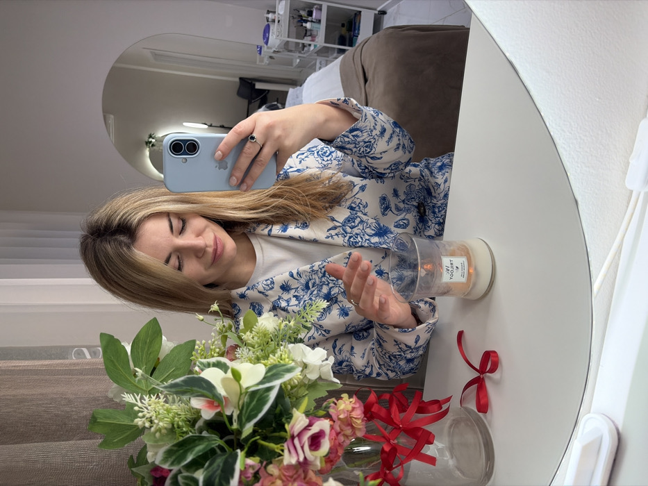 A blonde woman is captured in a mirror selfie within a beauty salon in Budva, Montenegro. She wears a white blazer with a blue floral pattern over a white ribbed top. Her right hand holds a light blue iPhone, pointed towards her, while her left hand is positioned near a clear glass candle jar containing an LED flickering light. On the white reflective surface foregrounding the woman, a clear glass vase holds a bouquet of pink, white, and green artificial flowers, secured with red ribbons. A loose red ribbon bow rests beside the vase. The background, visible in the mirror's reflection, depicts a professional treatment room featuring a light-colored treatment bed with a brown blanket, a white multi-tiered utility cart stocked with various beauty products, an arched wall mirror, and a standing ring light.