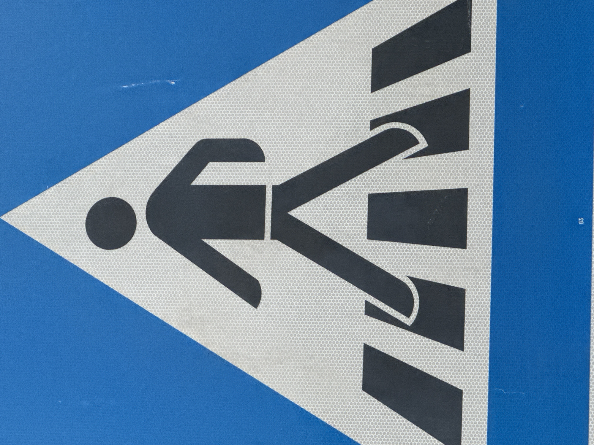 This is a close-up, straight-on photograph of a pedestrian crossing sign. The sign is triangular with a white, reflective interior on a bright blue background. In the center of the triangle, a black silhouette of a person is depicted walking across a black and white striped crosswalk. The person is shown with one leg forward and the other trailing, arms bent at the elbows. The crosswalk is represented by thick horizontal black stripes alternating with white spaces. The bottom of the sign has a solid blue rectangular section. In the lower right corner of the blue rectangular section, the small white numeral "03" is visible. There are minor scuffs and marks on the blue surface of the sign. The location is Unknown, Unknown.