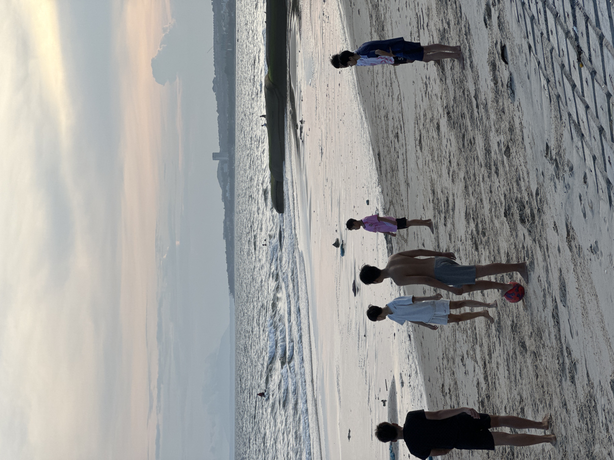 The image depicts a beach scene in Mui Ne, Vietnam. Several young males are present on the sandy shore, with some engaged in playing with a red soccer ball. The ocean with waves is visible in the background, and a distant coastline with buildings and hills suggests an urban or semi-urban setting. The sky is overcast with soft, diffused light, indicating either early morning or late afternoon. The overall atmosphere is relaxed and recreational, with the individuals appearing to be enjoying their time by the sea.