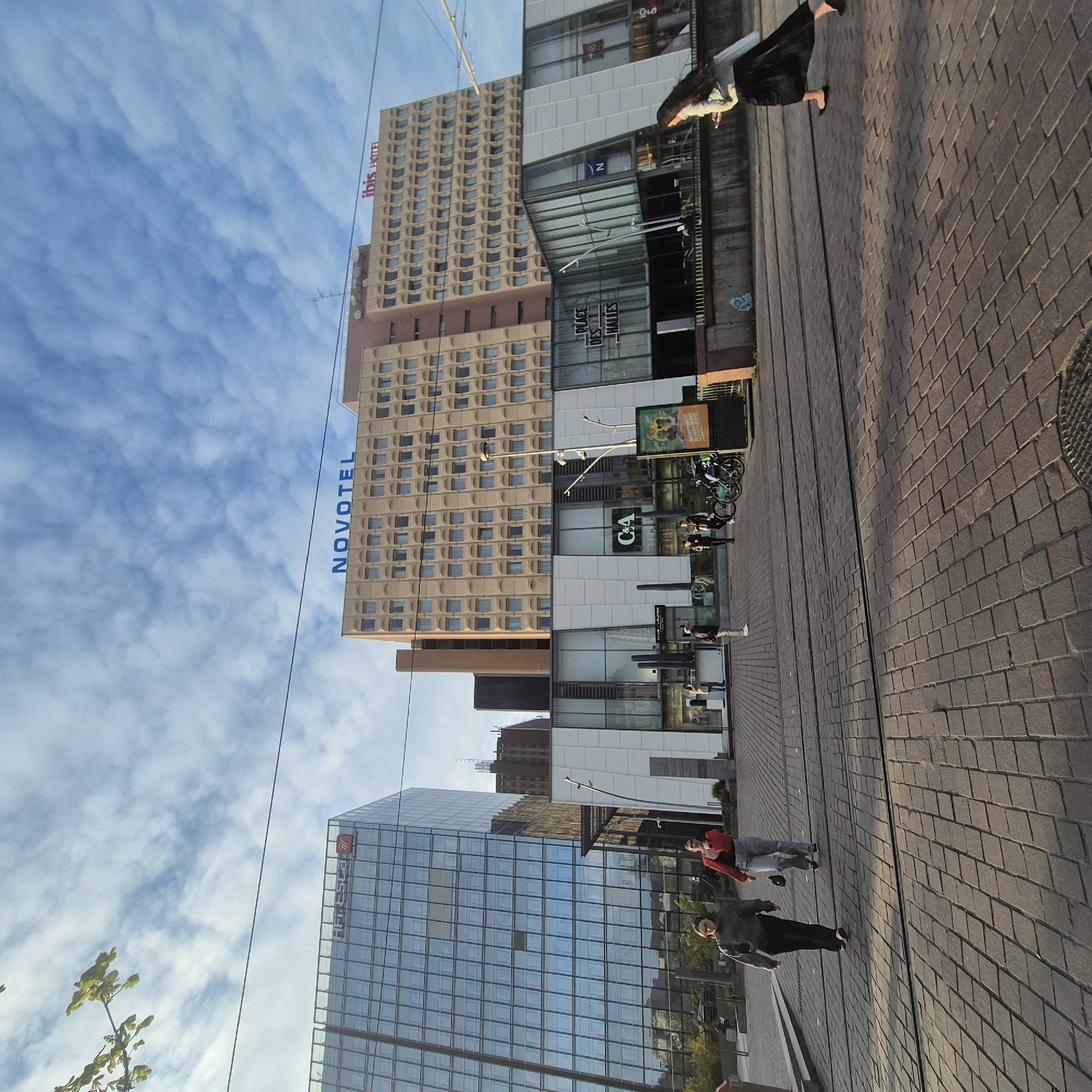 The image shows a street scene in Strasbourg, France. Several buildings are visible, including the Ibis Hotel, the Novotel, and another building with a mirrored facade bearing the name "PFI ESTEL". There is also a "C&A" store. People are walking along the brick-paved street. Bicycles are parked near the "C&A" store. The sky is partly cloudy.