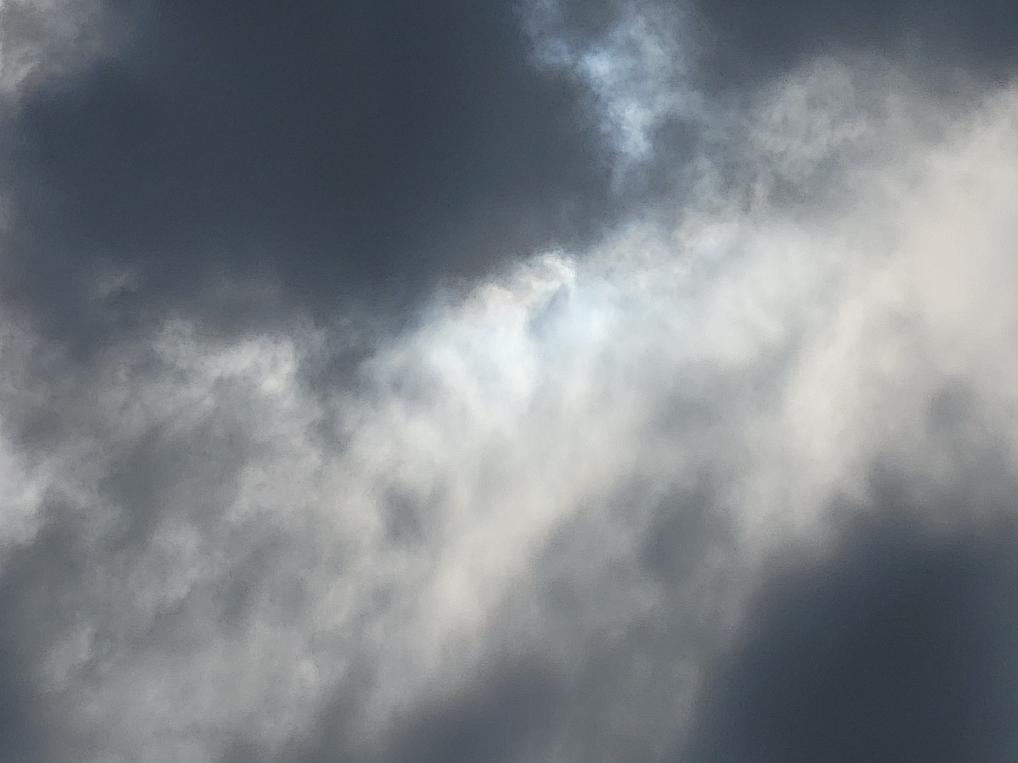 The image is a close-up shot of a cloudy sky. The clouds are dark and ominous in some areas, with lighter, more diffused clouds in others. The lighting suggests it might be overcast or a storm is approaching. There are no discernible landmarks or city features visible to indicate a specific location in Unknown, Unknown.