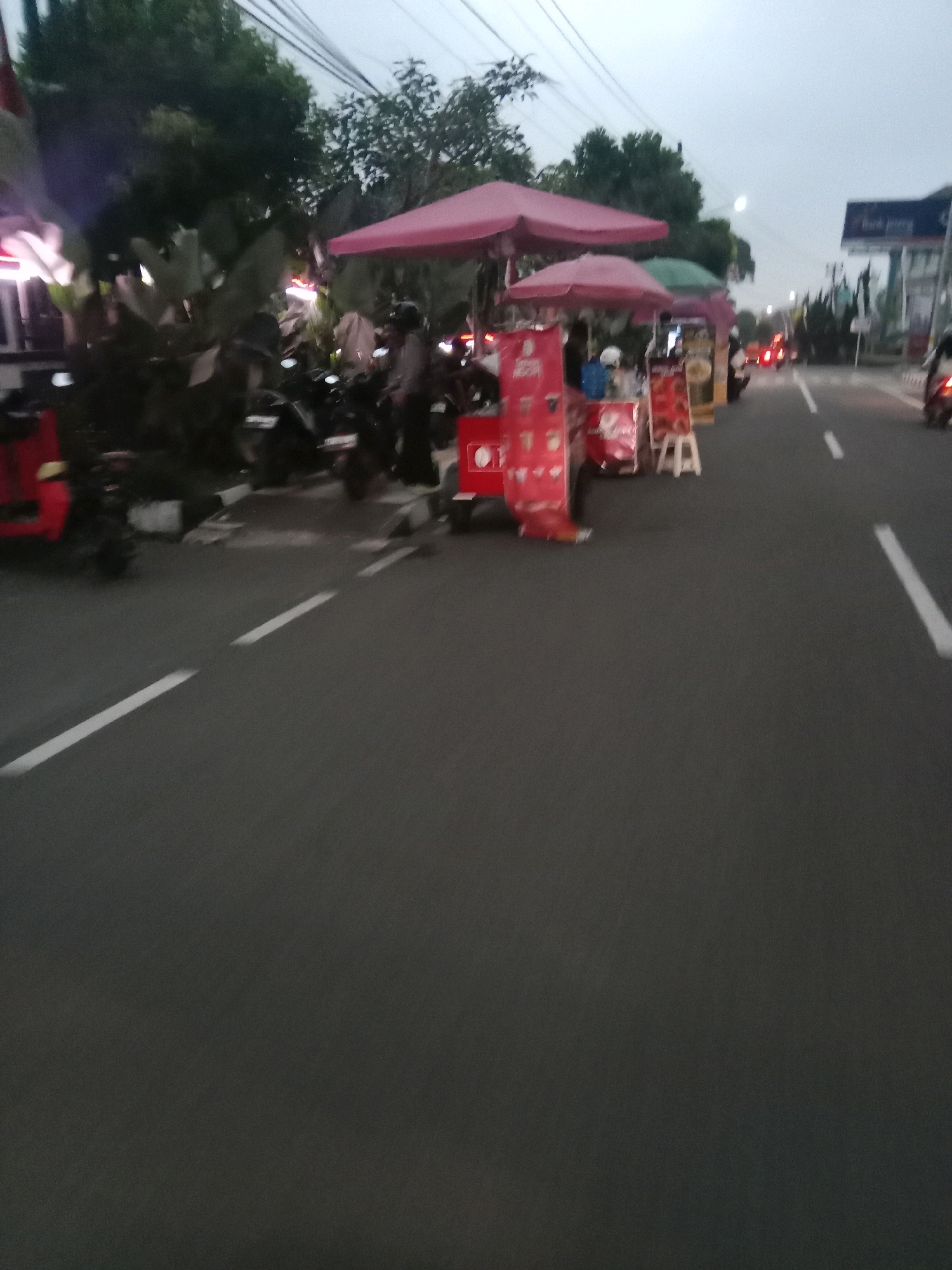 The image depicts a street scene in the city of Wonosobo, Indonesia, likely during twilight or early evening. The perspective is from a moving vehicle, as indicated by the blurred motion of the road and surrounding elements. On the left side of the road, several food stalls with red awnings and promotional banners are set up. These stalls appear to be selling beverages or snacks, with some featuring images of drinks. A few motorcycles are parked behind the stalls. Further down the road, more stalls and traffic lights are visible. On the right side of the road, a white line marks the lane for vehicles. Traffic is present, with some cars and motorcycles visible in the distance. The sky is overcast, contributing to the dim lighting conditions. The overall scene suggests a bustling street with commercial activity.