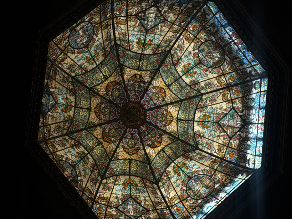 The image presents an upward-looking view of a large, elaborate octagonal stained-glass dome, situated in the Galería Güemes, located at Florida 165, between Avenida Corrientes and Avenida Rivadavia, in downtown Buenos Aires, Argentina.
The dome is constructed from numerous individual pieces of colored glass connected by a dark, intricate metal framework. The central element is a circular rosette with complex geometric and stylized floral motifs in reds, blues, and amber tones. Eight primary segments radiate outwards from this central point. Each segment is further subdivided into panels containing detailed imagery. Prominent features include large curvilinear panels adorned with vibrant floral garlands, ribbons, and foliage rendered in shades of green, blue, pink, and orange against a pale yellow and amber background. Interspersed within these radiating segments are circular medallions, each depicting allegorical or classical figures, such as nymphs or cherubs, engaged in various poses, rendered in muted tones of white, beige, and light blue. The overall color palette is rich, dominated by amber, emerald green, sky blue, rose pink, and deep red, creating a harmonious mosaic. The dome is illuminated from above, indicating it serves as a skylight, with natural light passing through the colored glass. The edges of the dome are bordered by a dark, possibly ornate, architectural trim, consistent with interior ceiling ornamentation. No people are visible within the frame; the scene is static, focused solely on the architectural and artistic details of the dome.