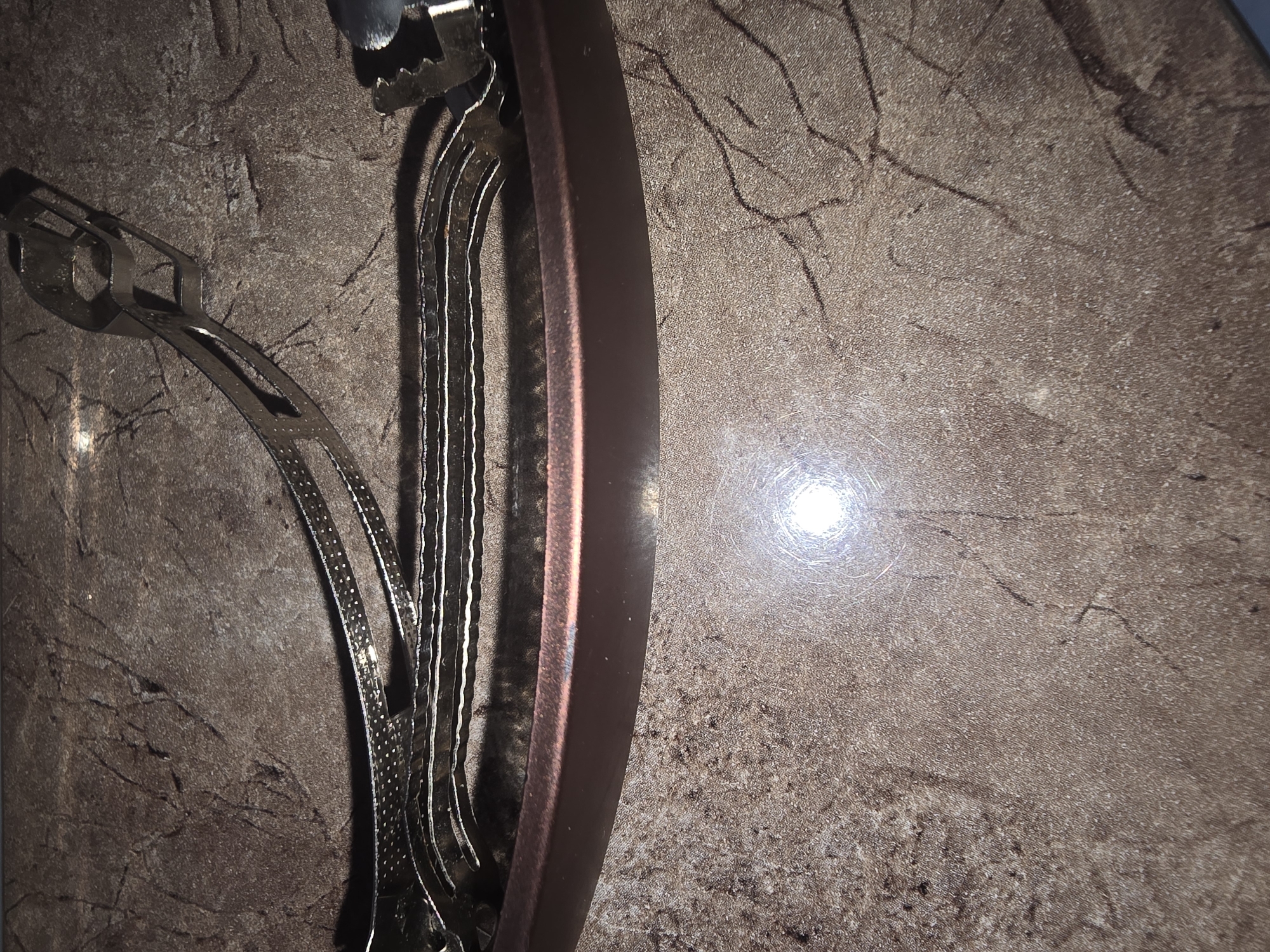 The image shows a close-up, high-angle view of a hair clip lying on a mottled brown surface, possibly a countertop or table. The hair clip is silver-toned metal with a slightly curved design, featuring a series of layered metal strips and a hinge mechanism at one end. The surface it rests on has a textured pattern with darker lines resembling cracks or veins. A bright light source is reflected prominently on the surface, creating a circular glare in the lower portion of the image. There is no indication of the city of Ulan-Ude, Russia, or any people in the image.