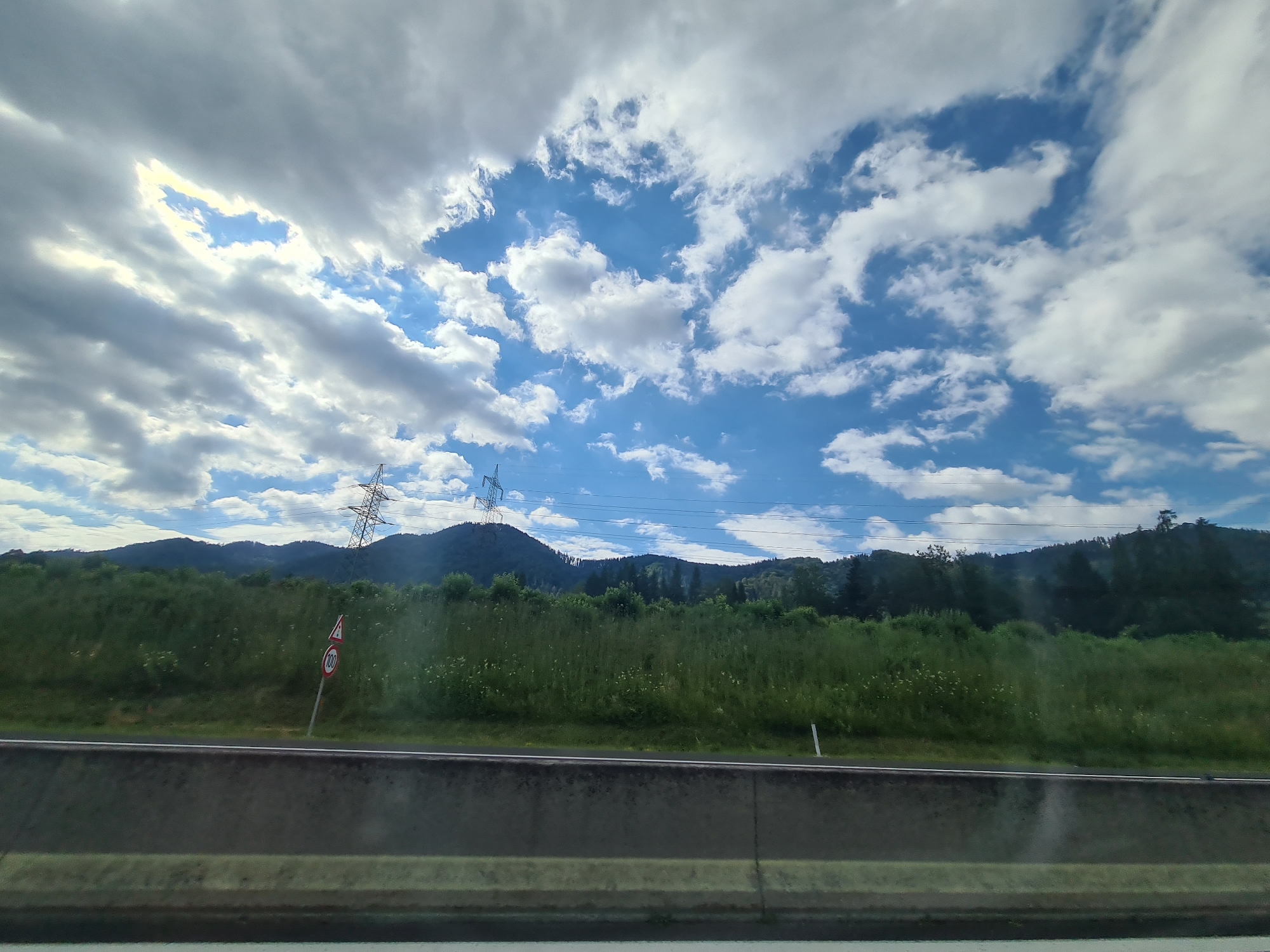 This is a photograph taken from a moving vehicle on a highway in Krieglach, Austria. The image captures a landscape scene with a cloudy sky, green hills, and trees. In the foreground, a concrete barrier runs along the side of the highway. To the left, a traffic sign indicating a speed limit of 100 is visible, partially obscured by grass. Further back, two large metal pylons stand against the backdrop of the hills. The sky is a mix of blue patches and white, fluffy clouds, with some areas of sunlight breaking through. The overall impression is one of a scenic drive through the Austrian countryside.