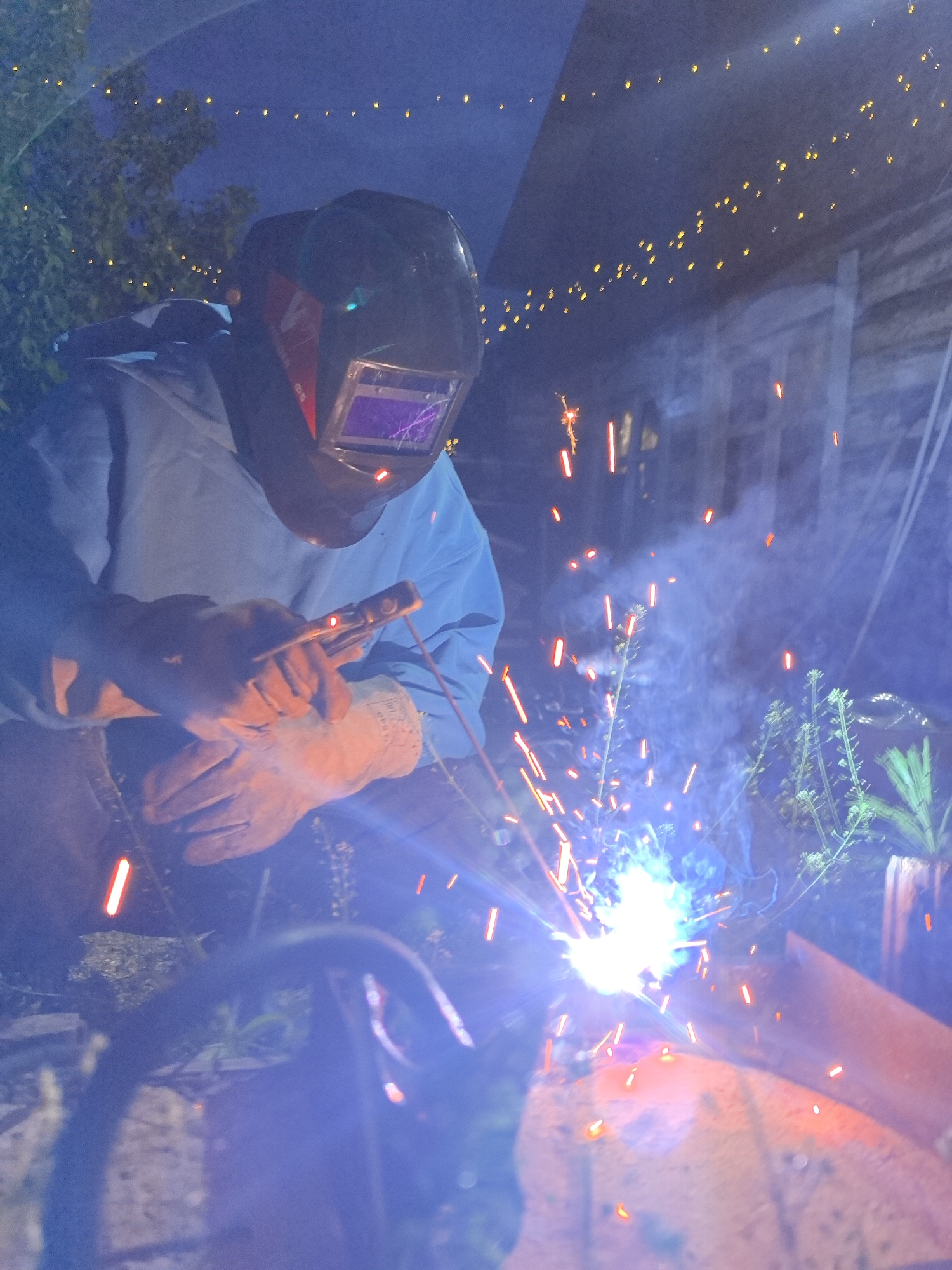 A person wearing a welding mask and gloves is welding metal. Sparks fly from the welding point. There are plants visible in the background. Overhead, there are string lights. The location of Noviy, Russia, is not confirmed.