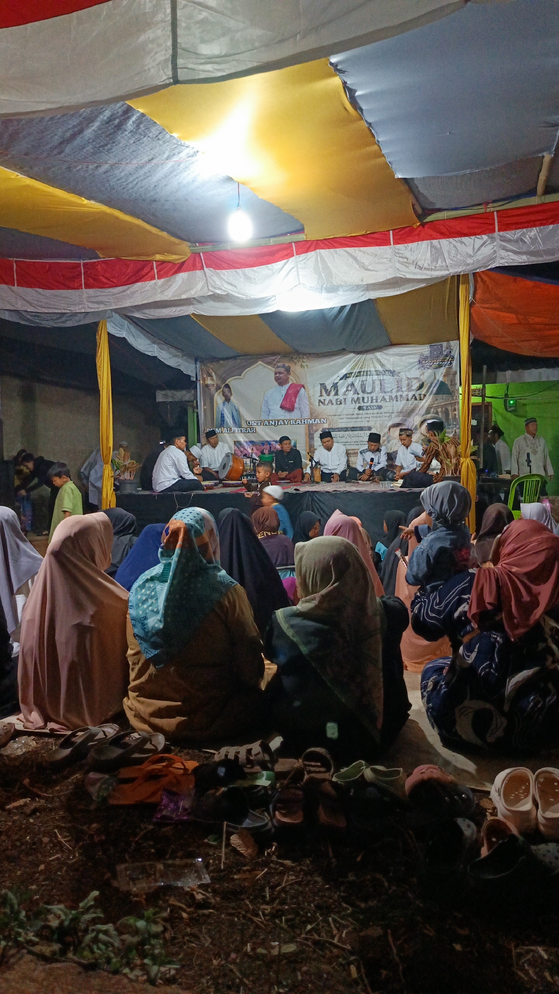 An outdoor evening gathering is taking place under a makeshift tent structure in Margajaya, Indonesia. The tent is supported by poles and draped with fabric in red, white, yellow, and grey. A bright bare bulb hangs from the ceiling, illuminating the scene.
On a raised platform at the back, several individuals are seated, likely performers or speakers. A large banner behind them features images of men and text that appears to be religious in nature, mentioning "MAULID NABI MUHAMMAD SAW" and "UST. ANJAYRAHMAN". Some individuals on the platform are holding instruments, including what looks like a drum.
In front of the platform, a large audience, primarily women and children, are seated on the ground, facing the stage. Many are wearing hijabs and modest clothing. The ground appears to be dirt and covered with debris. A collection of footwear, including sandals and flip-flops, is scattered in the foreground, suggesting attendees removed their shoes before entering the gathering area. A few children are visible walking around the periphery of the audience. The overall atmosphere suggests a community event, possibly a religious celebration.