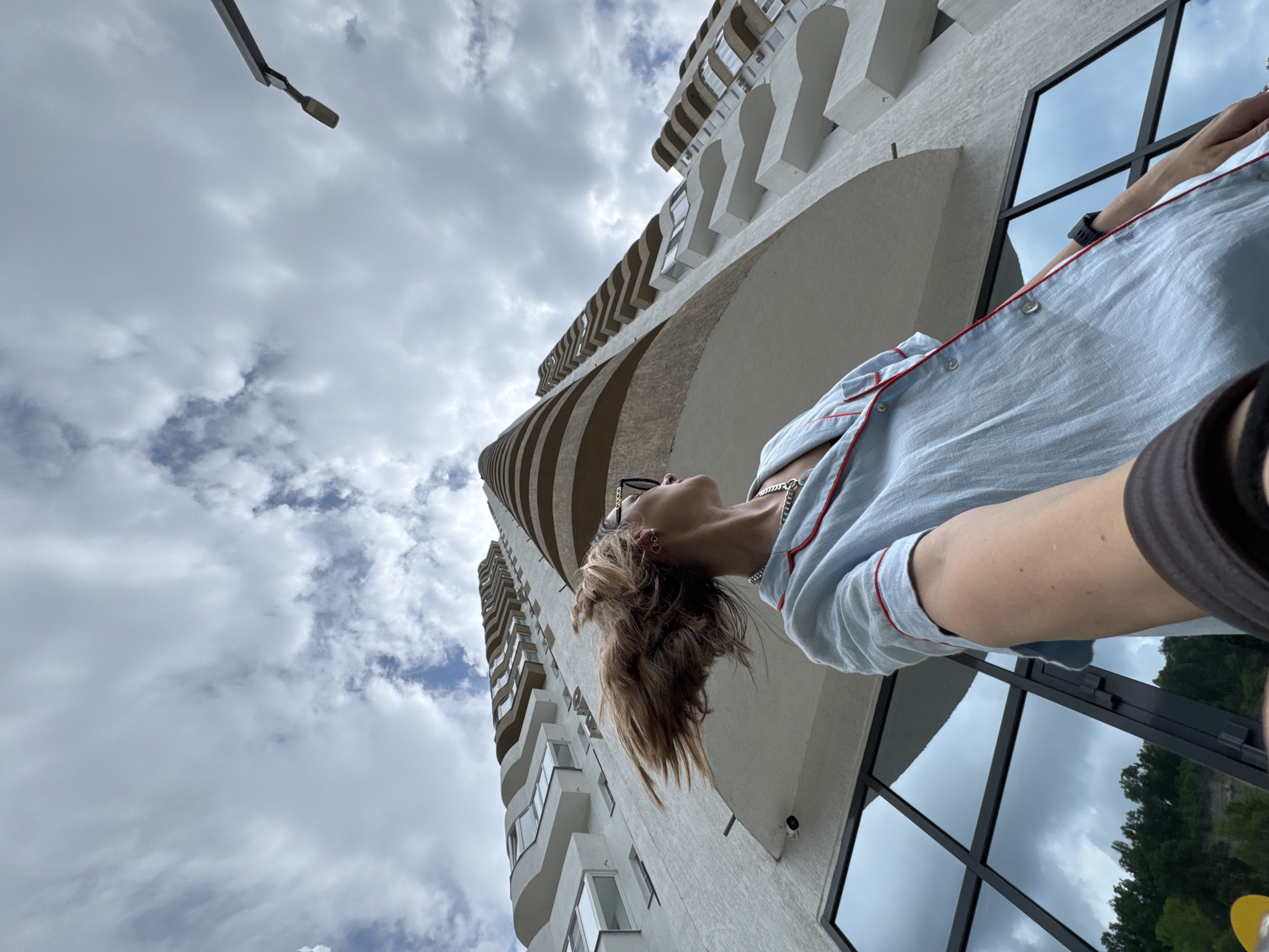 A low-angle, wide shot captures a person looking up at a modern, multi-story building with curved balconies against a cloudy sky. The building appears to be made of white concrete with dark-framed windows. In the foreground, the person's face is visible in profile, with their hair blowing to the left. They are wearing a light blue collared shirt with red trim and a silver chain necklace. Their right arm extends towards the camera, holding a dark object, possibly a camera or phone, suggesting a selfie. The reflection in the building's windows shows clouds and a treed hillside. A streetlamp is visible in the upper right corner. The scene is set in Sochi, Russia.